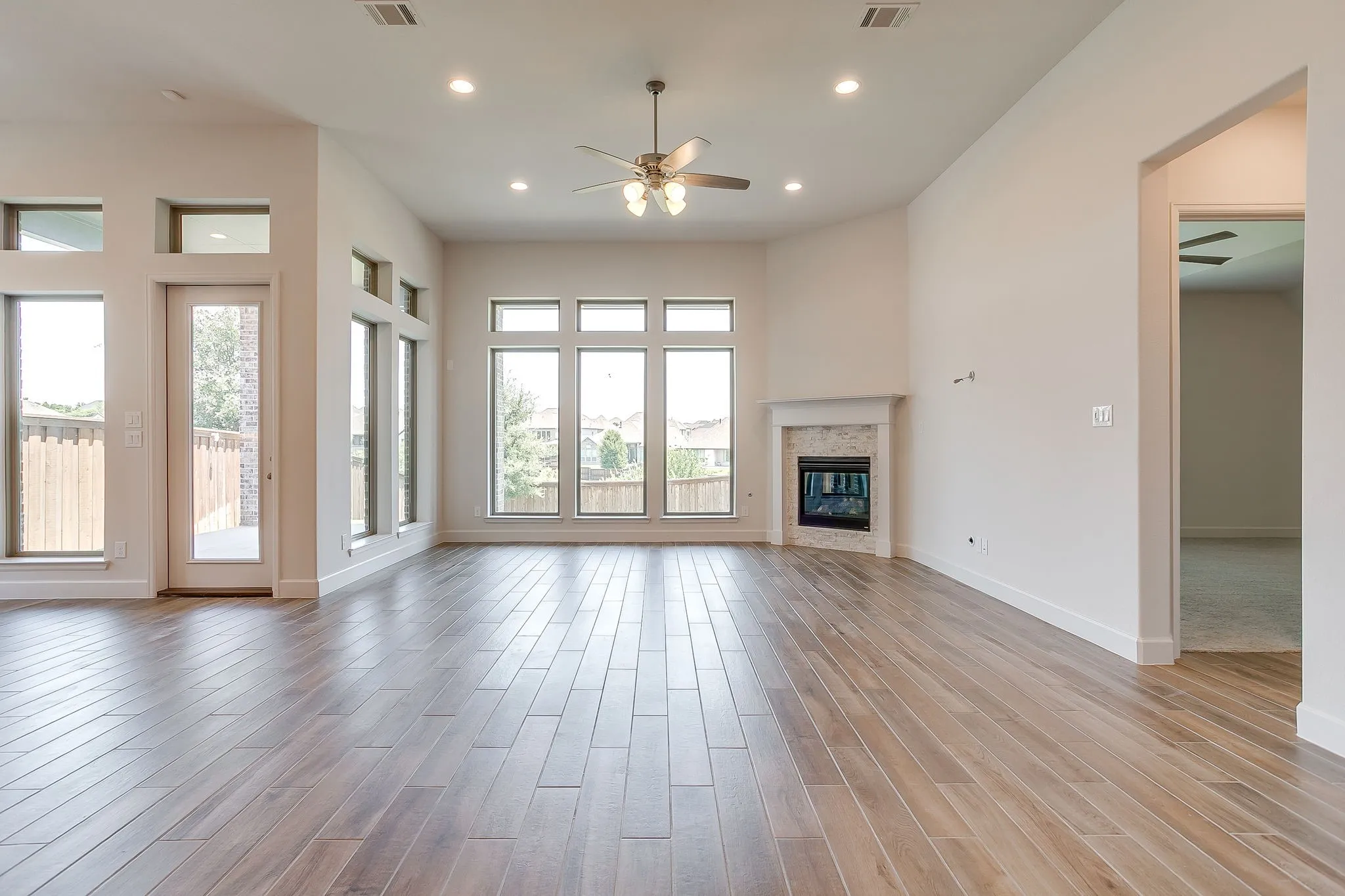 Expansive interior space featuring wood-finish flooring, a corner fireplace with stone surround, a ceiling fan, recessed lighting, and multiple large windows with transom details