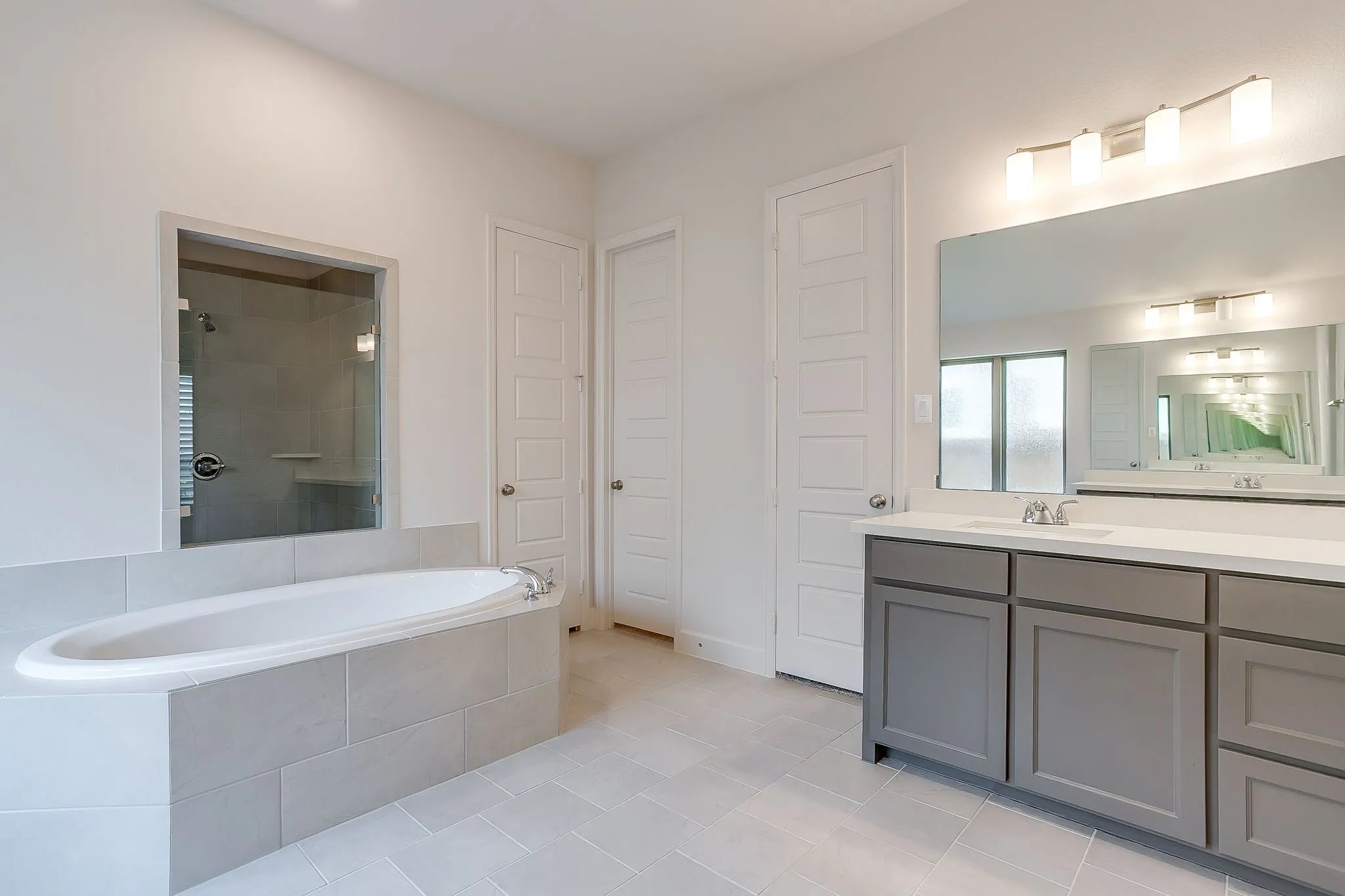 Spacious bathroom featuring a built-in soaking tub with tiled surround, a glass-enclosed shower, a double vanity with gray cabinetry and white countertops, and light-toned tile flooring