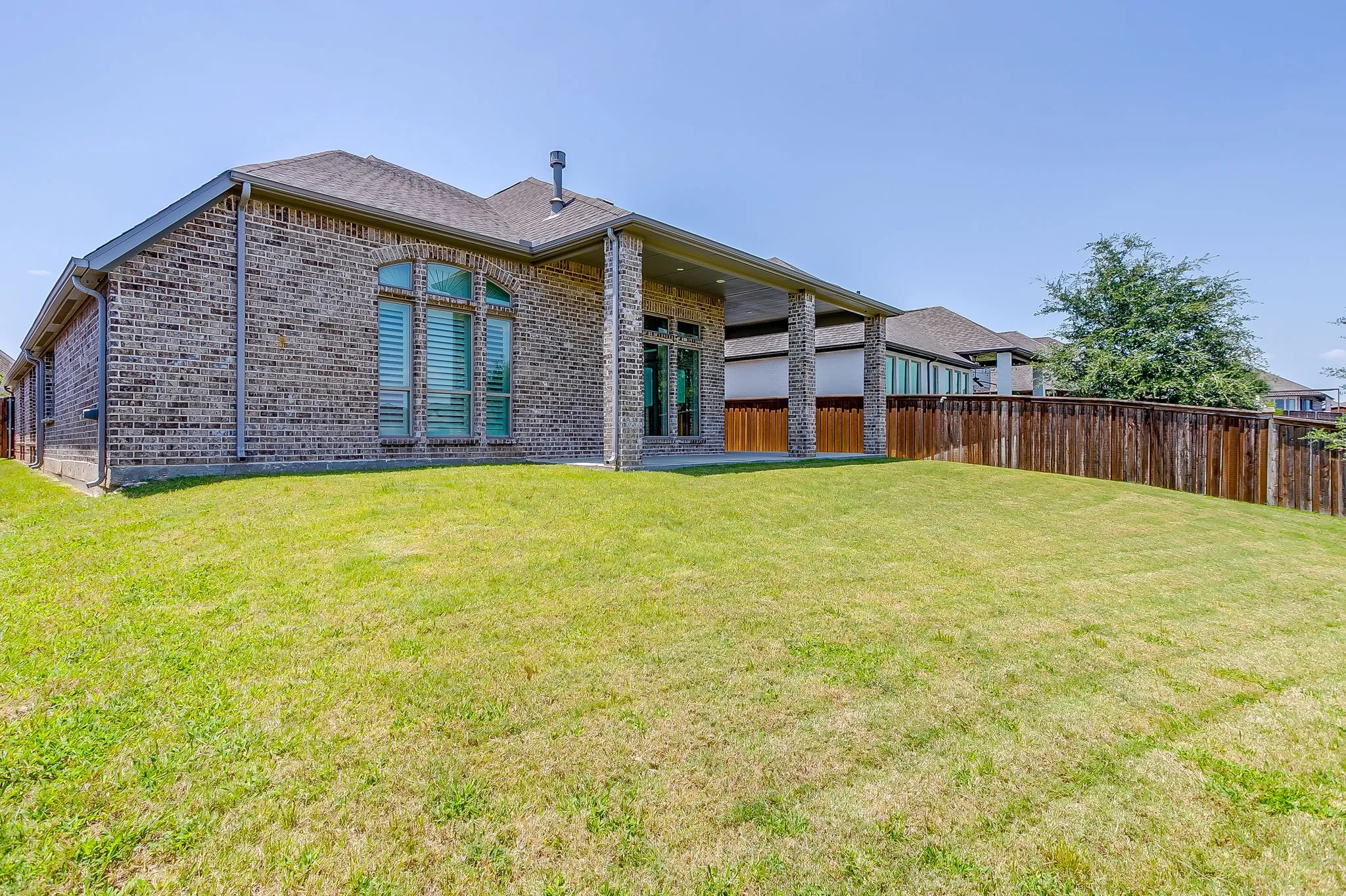 Expansive backyard featuring a manicured lawn, full privacy fencing, and a covered patio with stone-accented pillars