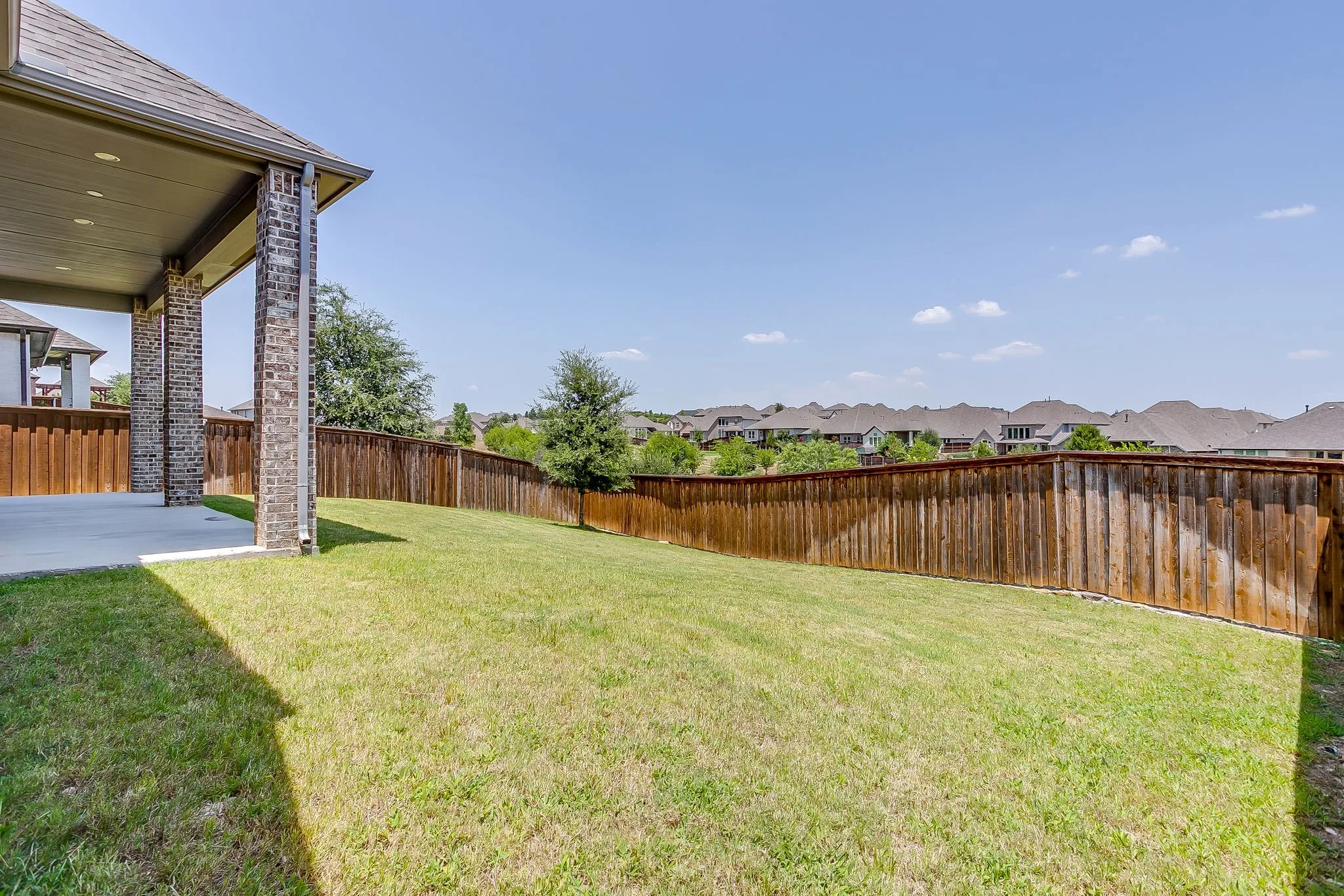 Expansive backyard featuring a covered concrete patio, lush green lawn, full wood fencing, and mature landscaping