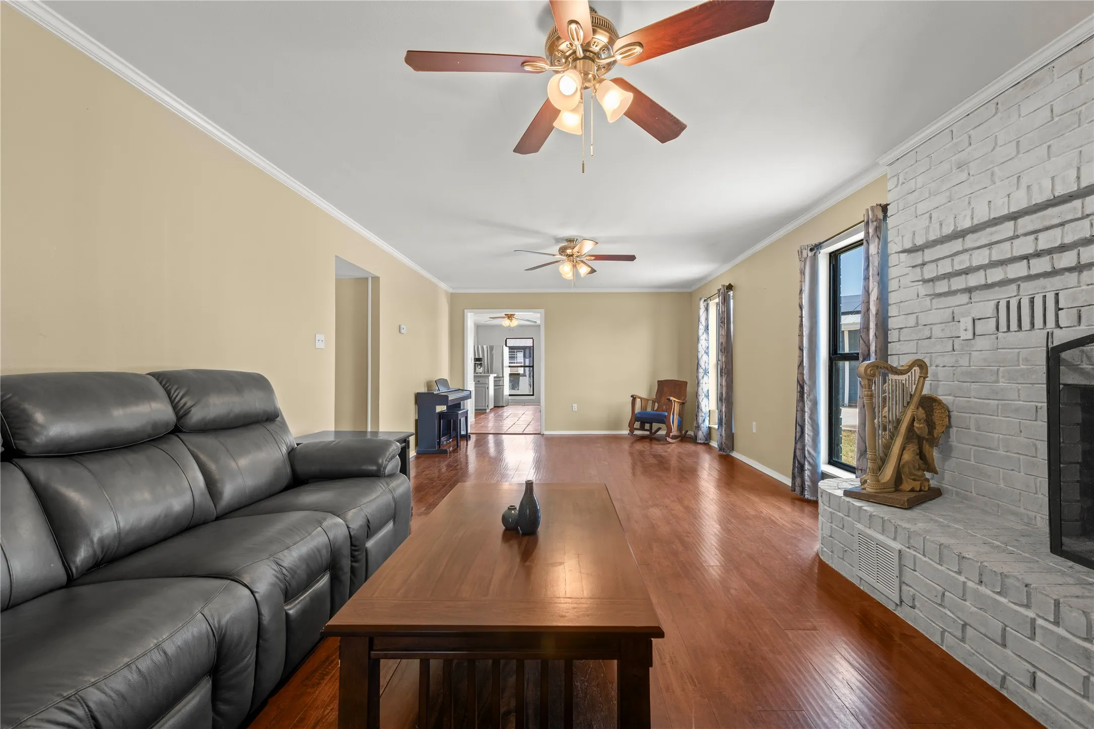 Spacious living area with wood floors, dual ceiling fans, and a classic painted brick fireplace, opening into the kitchen and dining spaces.