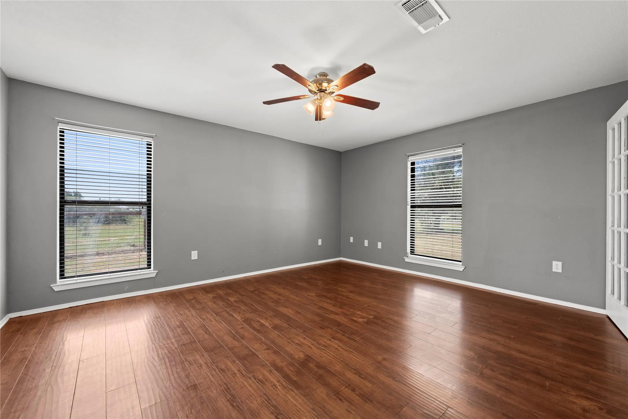 Spacious primary bedroom with rich wood flooring, gray walls, and abundant natural light from dual windows