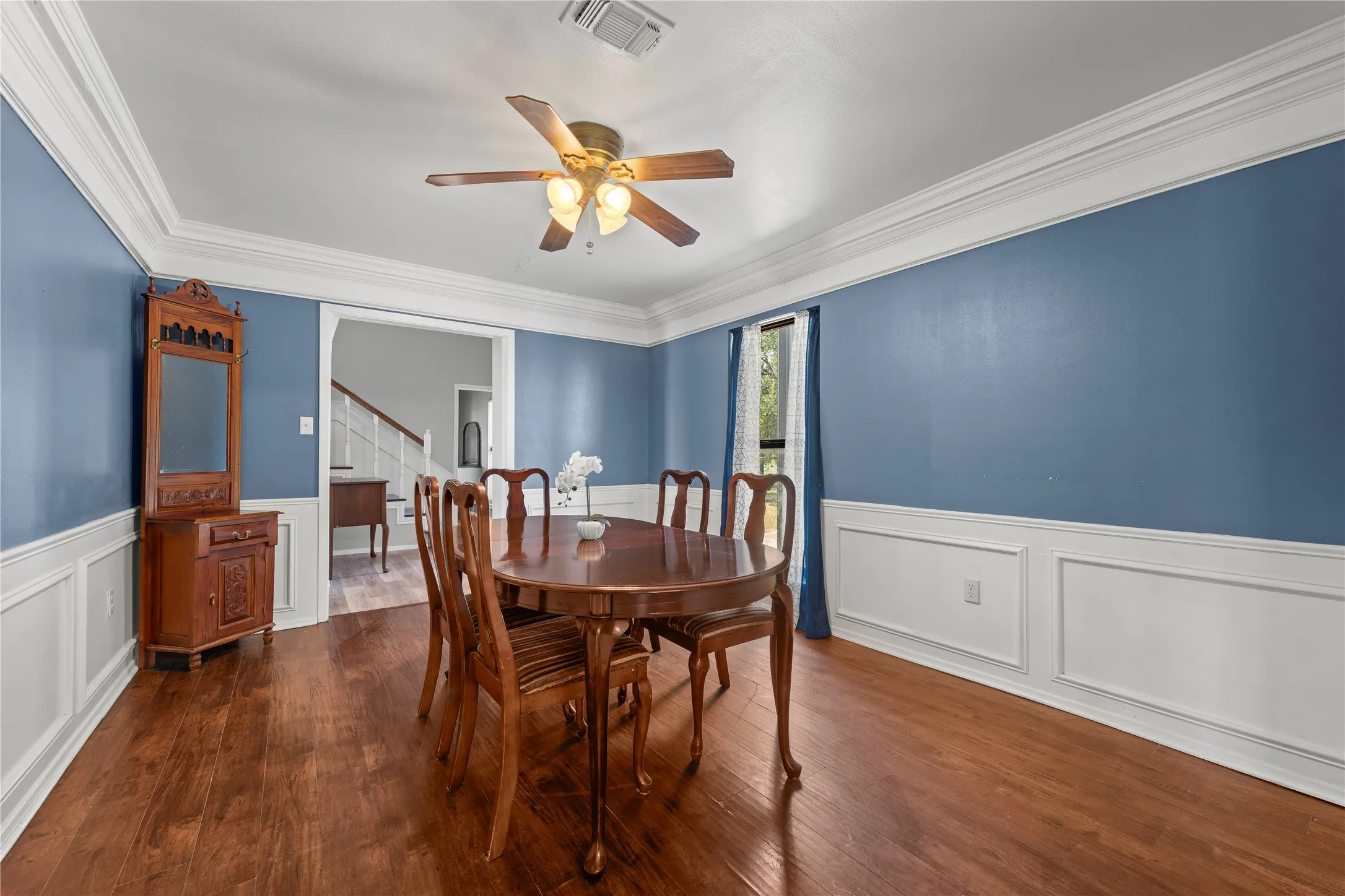 Elegant formal dining room featuring rich blue walls, crown molding, wainscoting, and a ceiling fan for year round comfort