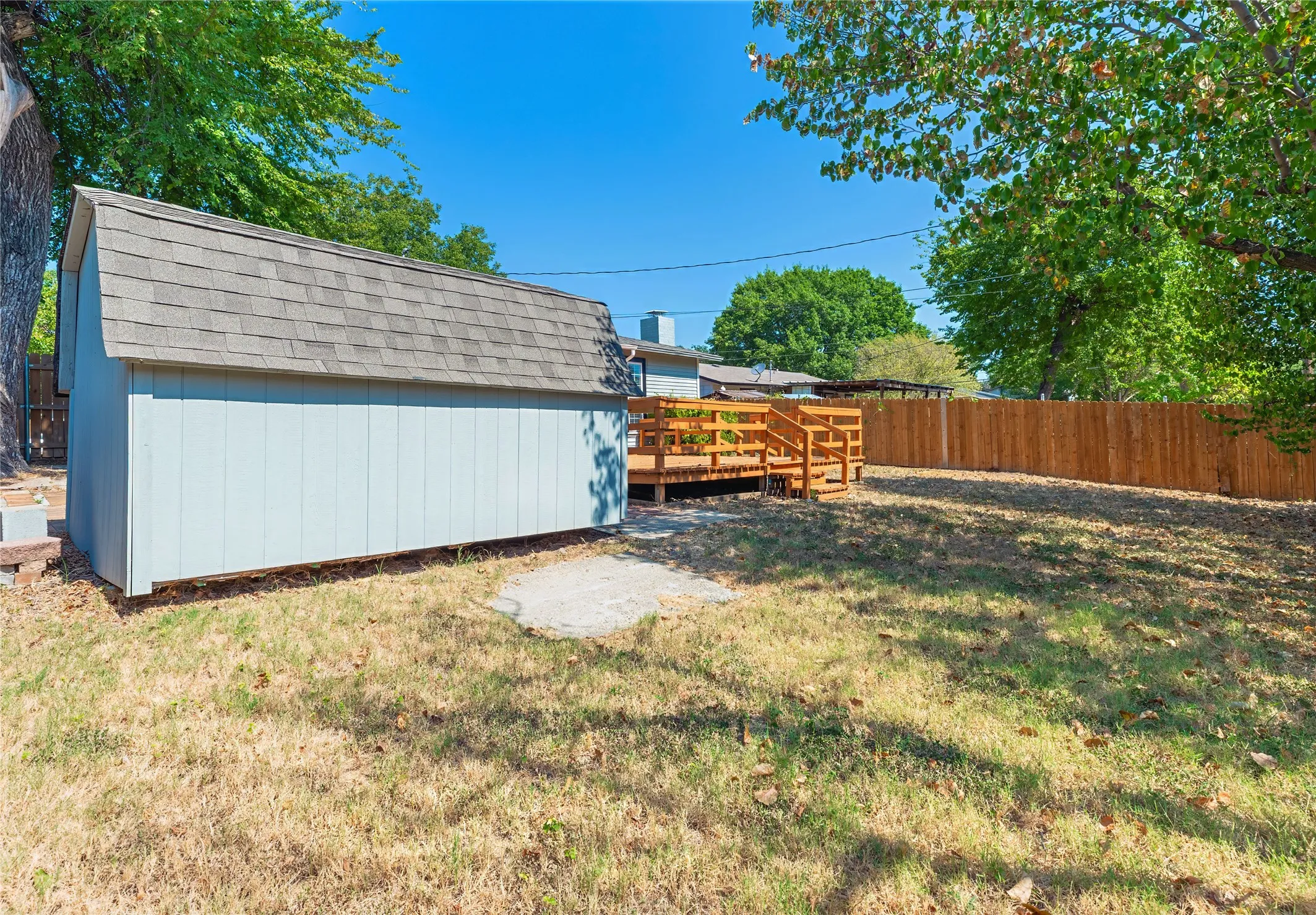 Detached storage shed with a gambrel roof and shingle siding, accompanied by a wood deck with railings and a wood privacy fence