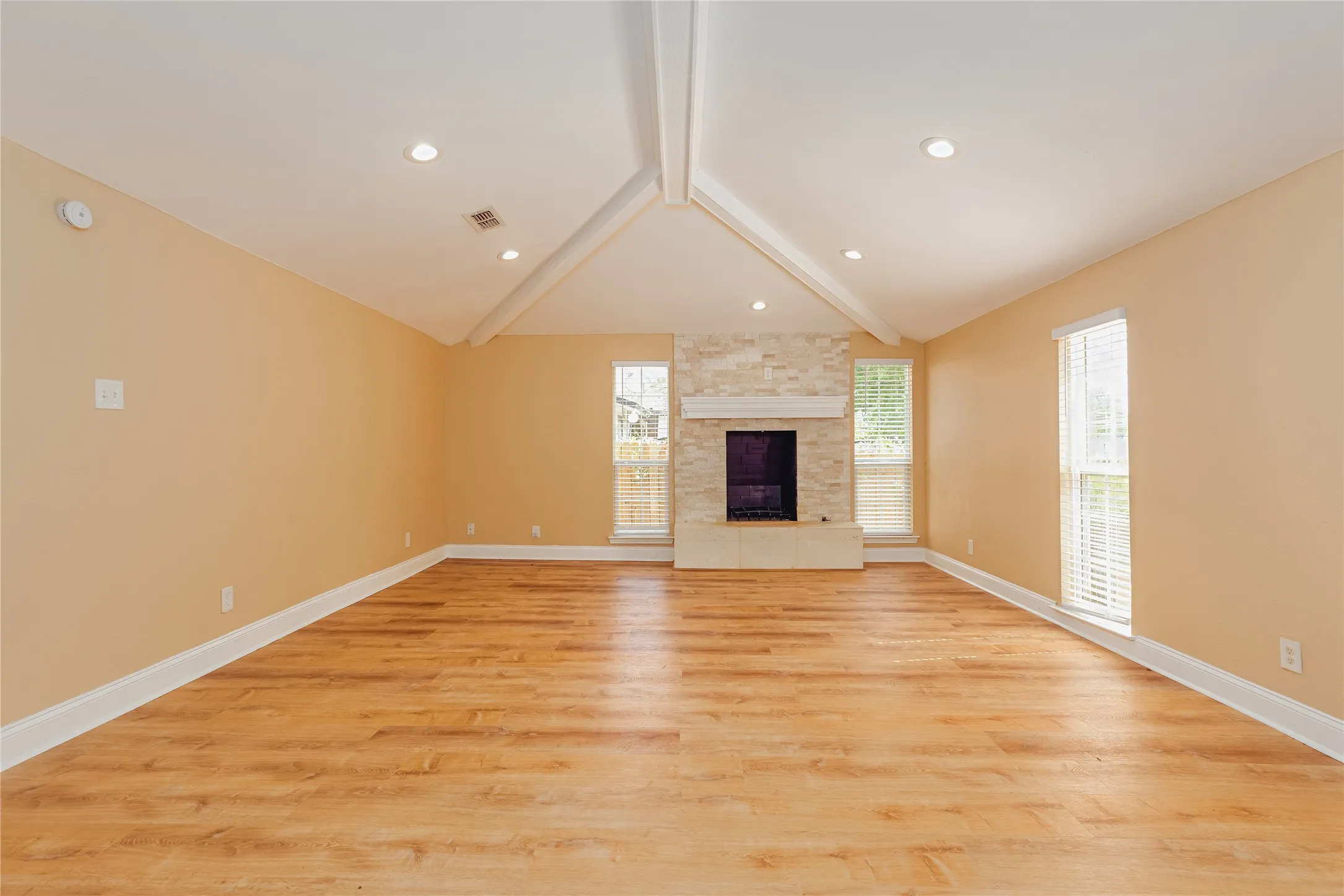 Vaulted ceiling with exposed beams, LVPh flooring, recessed lighting, stone-clad fireplace with mantel, and multiple windows with blinds