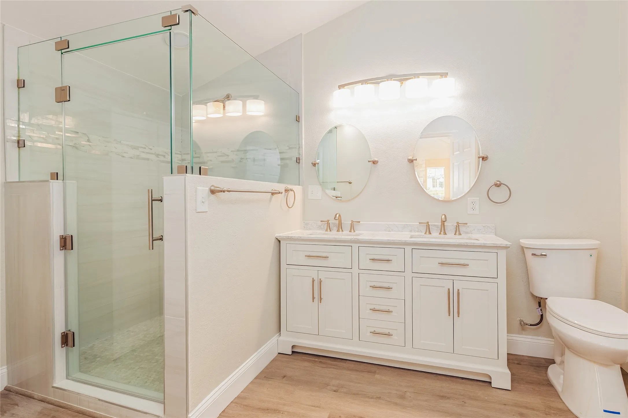 Walk-in shower with clear glass enclosure, dual vanity with white cabinetry, dual oval mirrors, and LVP flooring