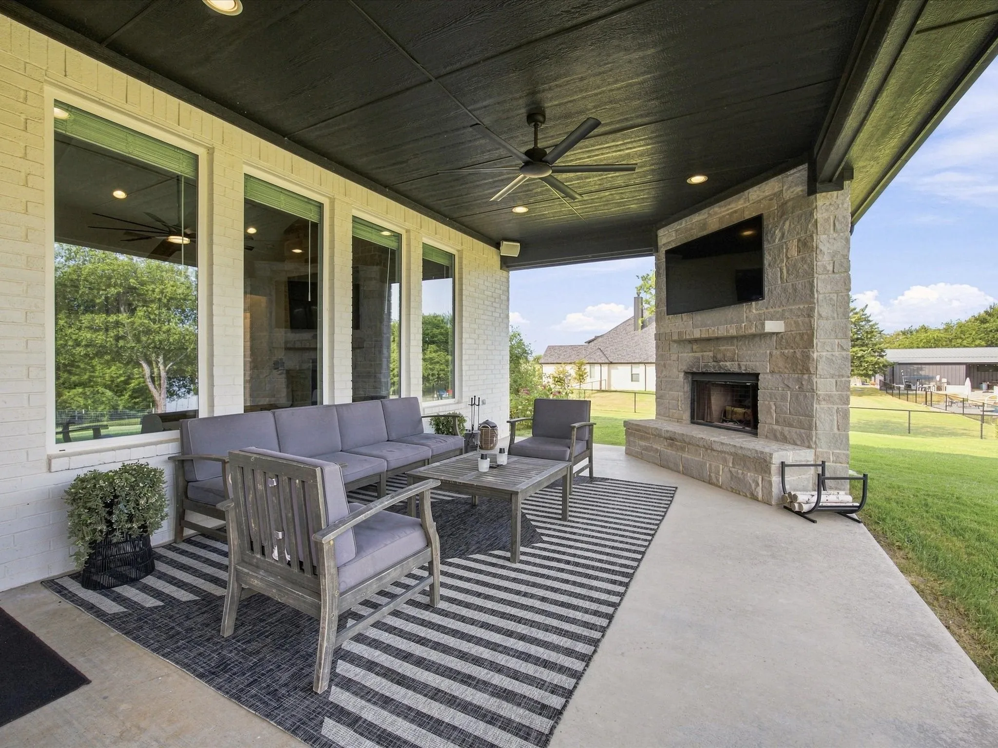 Expansive covered patio featuring a stone-clad fireplace, built-in television alcove, overhead ceiling fan, recessed lighting, and large windows