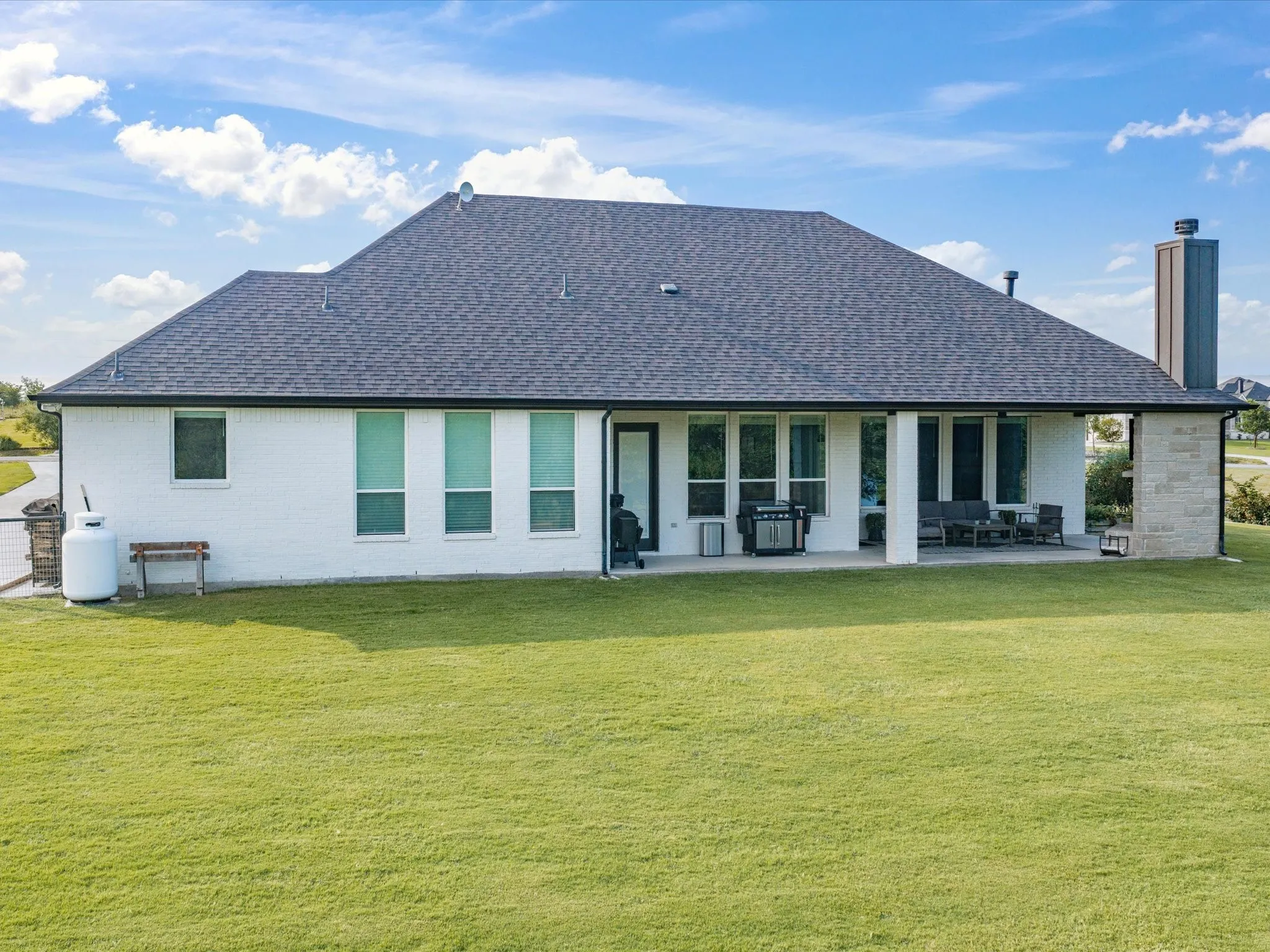 Expansive rear yard with a covered patio, white brick exterior, dark asphalt shingle roof, and a stone-clad chimney