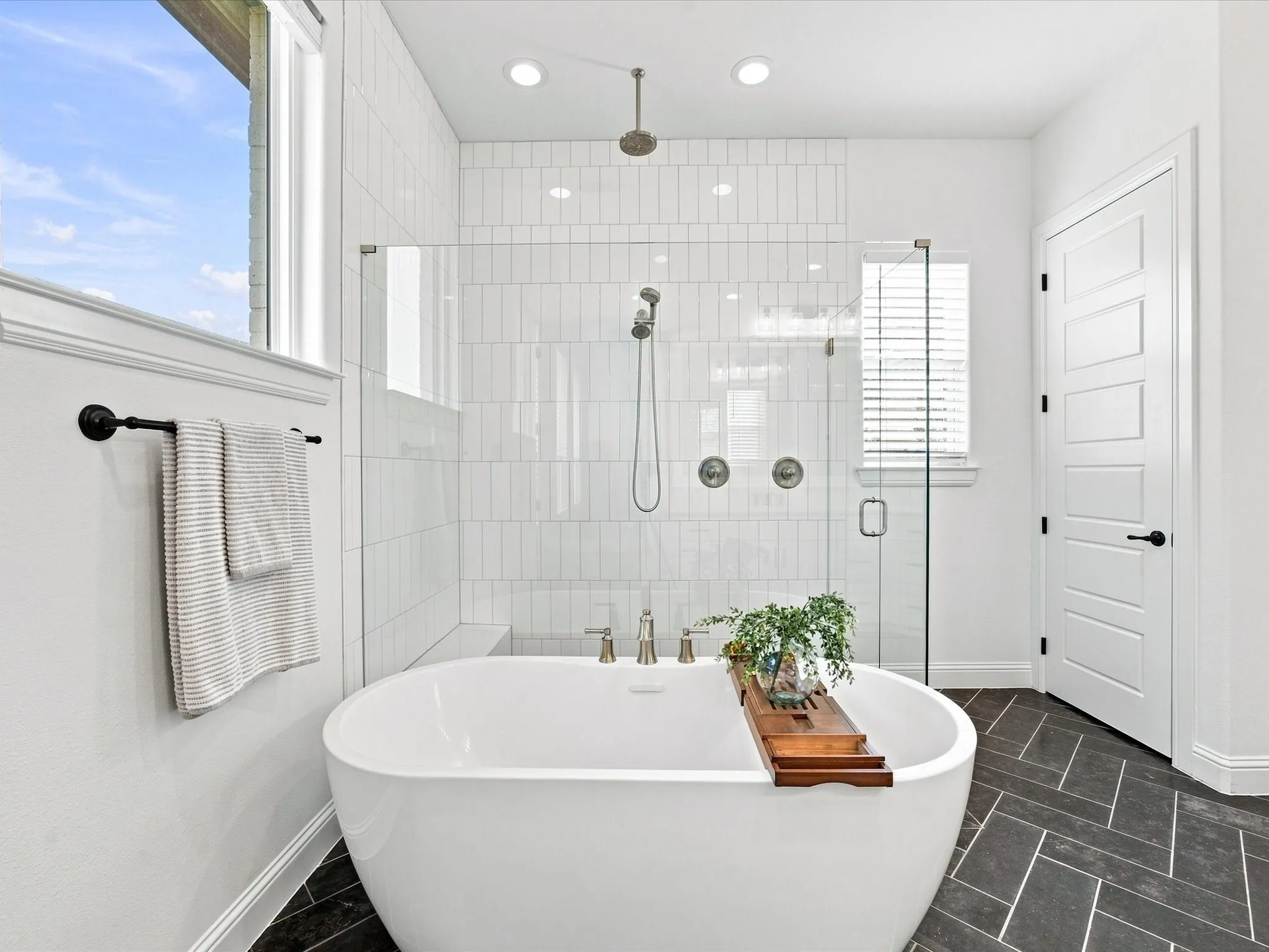Freestanding soaking tub, walk-in shower with vertical tile, glass shower enclosure, dark herringbone tile flooring, and a window with natural light