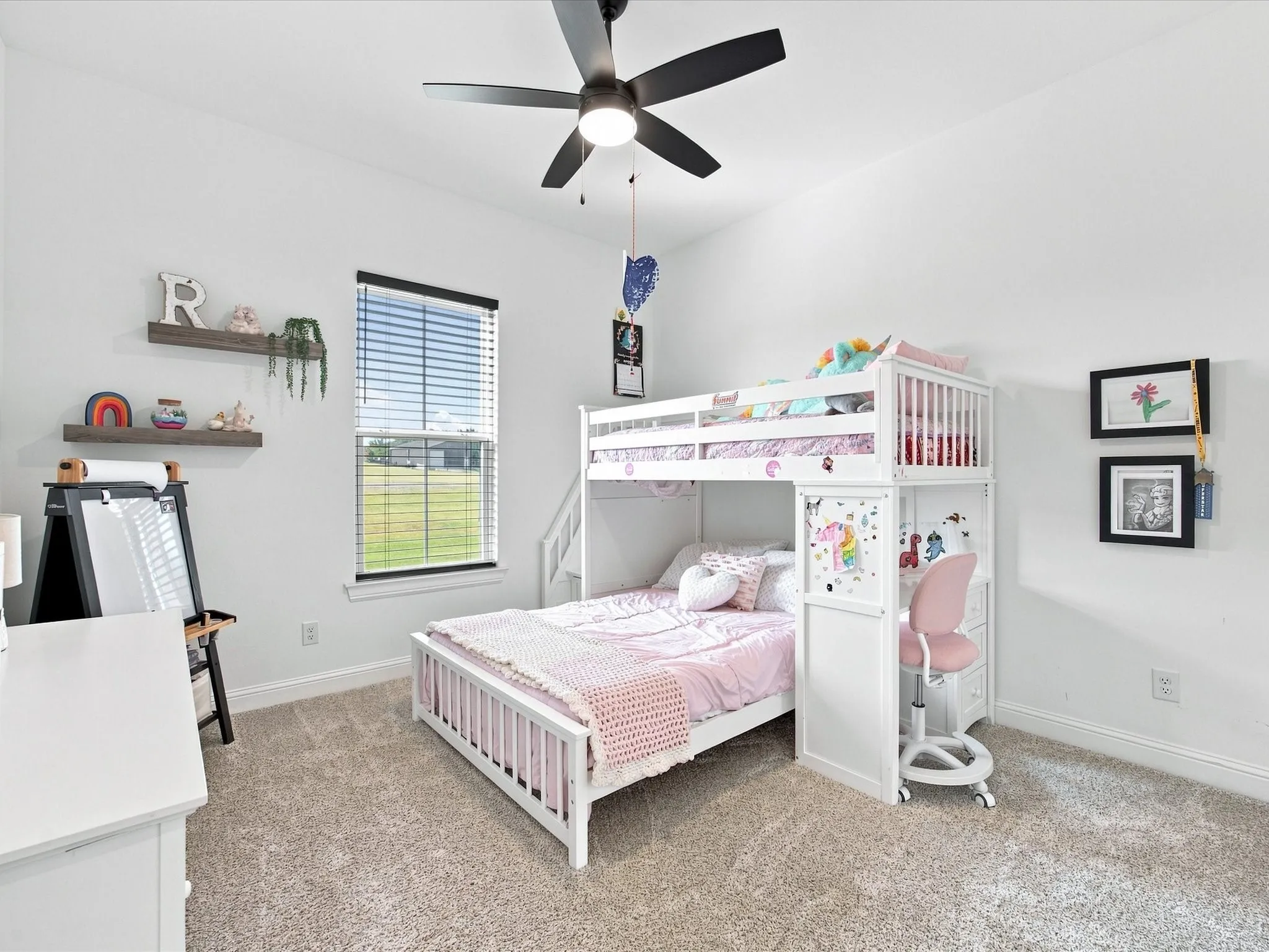 Bright bedroom featuring carpet flooring, a ceiling fan with integrated lighting, horizontal window blinds, white loft-style bunk bed with built-in storage and desk space, floating shelves, and framed wall-mounted artwork