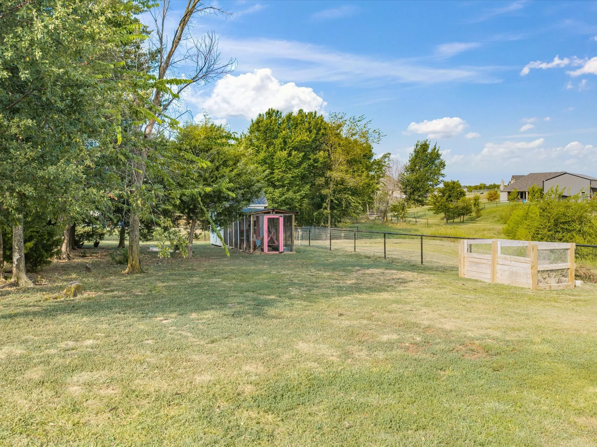 Expansive grounds featuring mature shade trees, manicured lawn, fenced pasture area with gated entry, detached storage structures, and pastoral backdrop with distant outbuildings