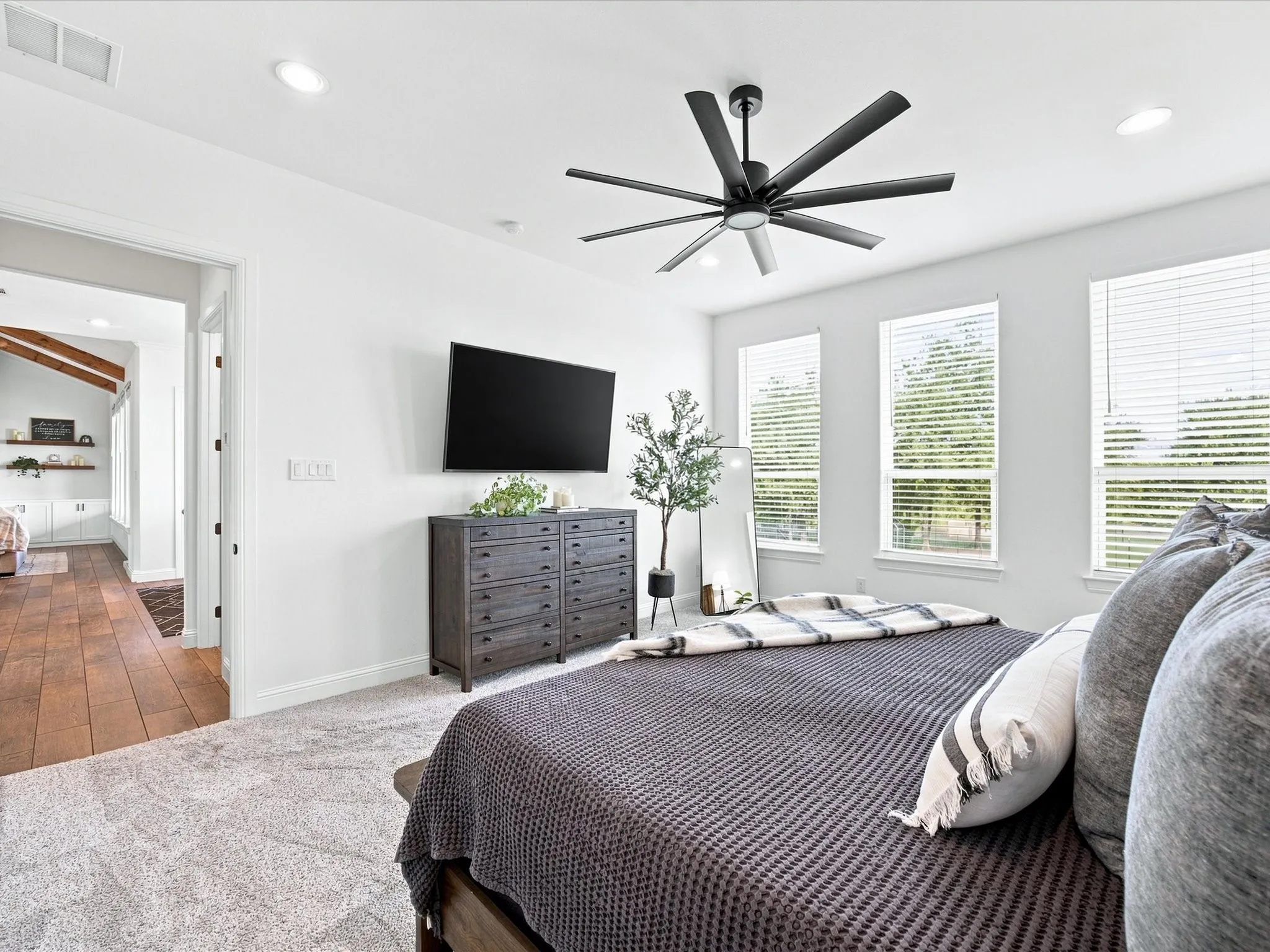 Primary bedroom suite showcasing a modern ceiling fan with dark metal finish, wall-mounted flat-screen television above a dark wood dresser with decorative décor, abundant natural light from multiple large windows with plantation shutters, and neutral wall-to-wall carpeting throughout