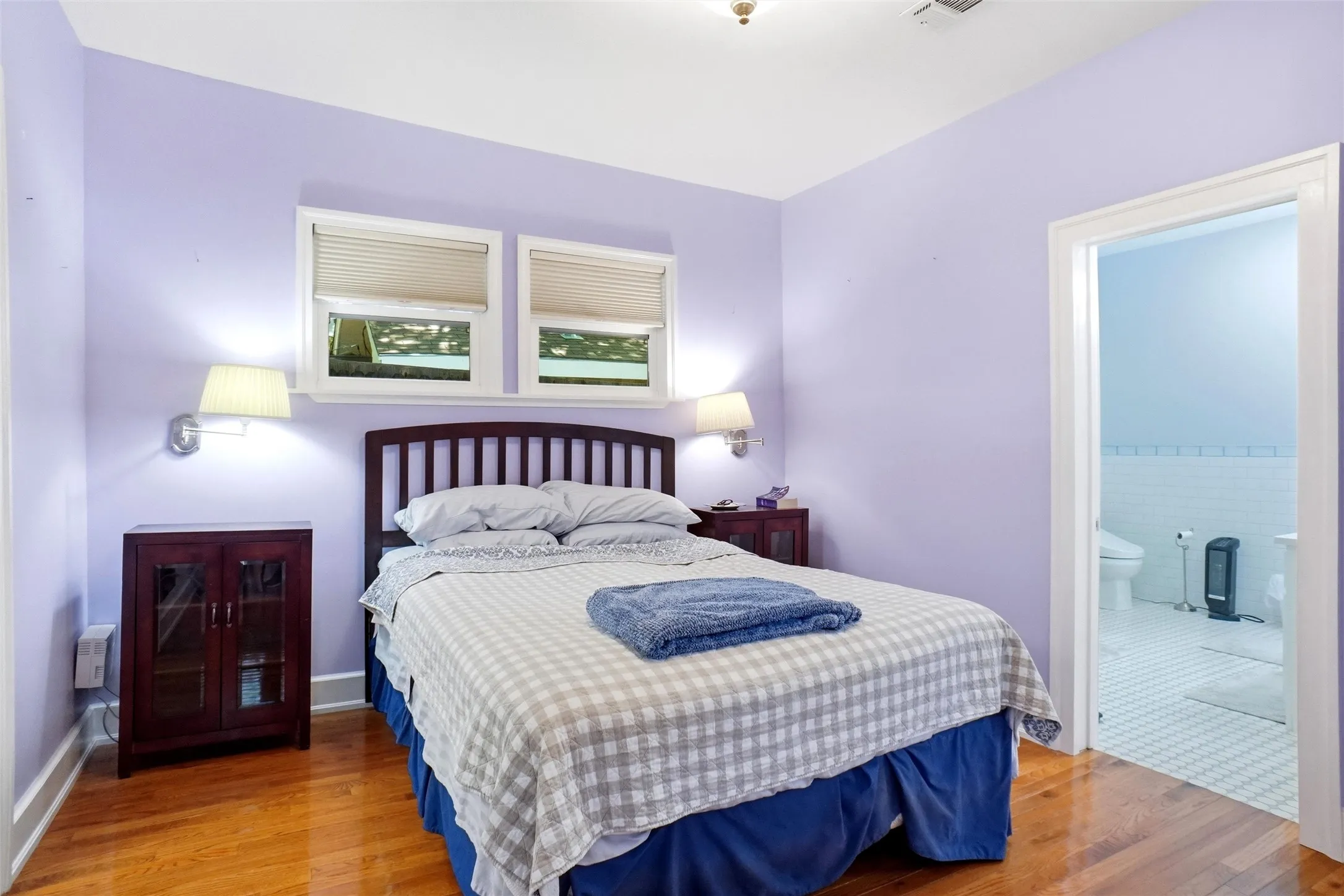 Hardwood flooring throughout this room featuring a wooden headboard, two sconce light fixtures, and windows with blinds