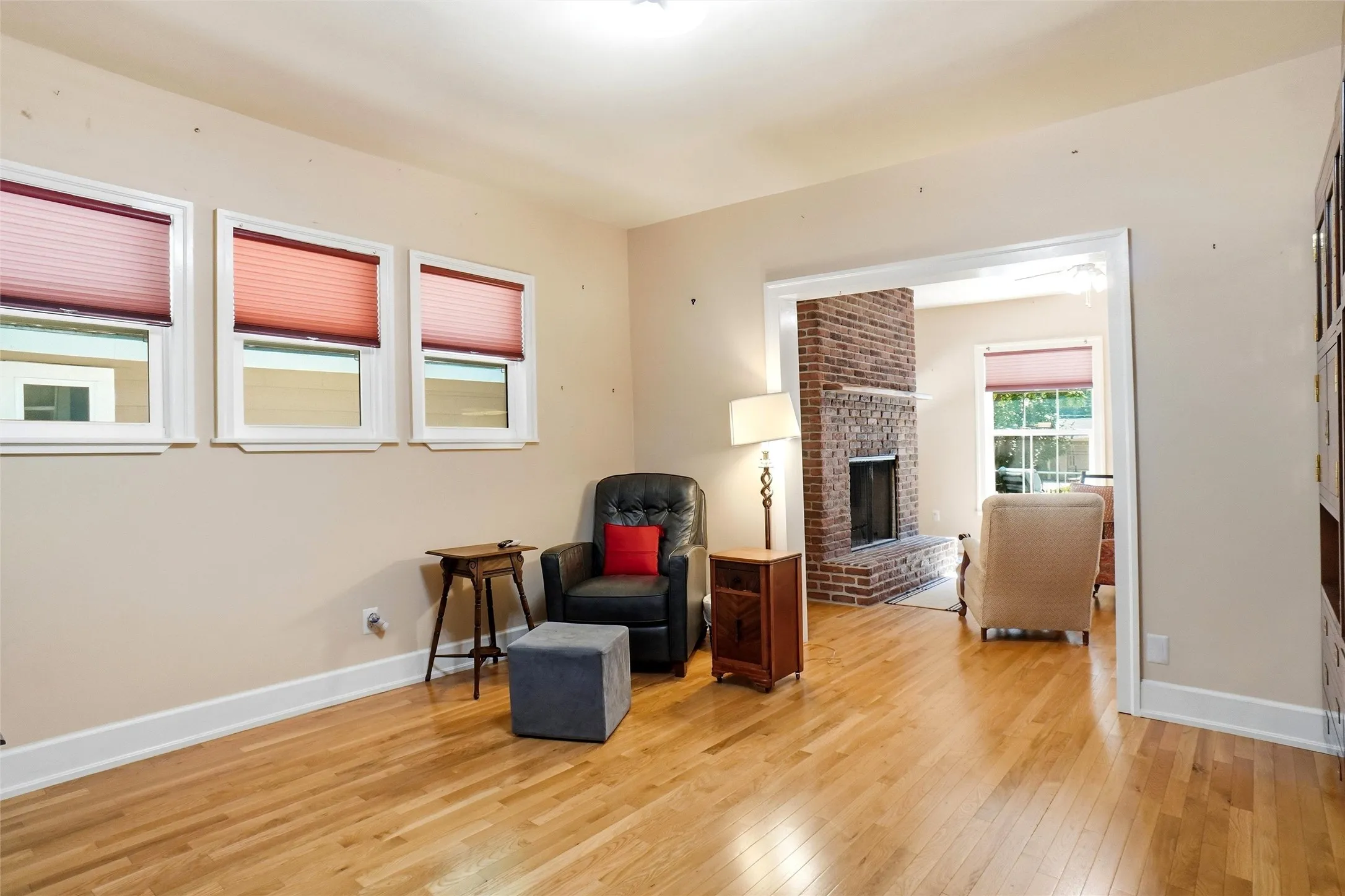 Hardwood flooring extends throughout this room, featuring three windows with white trim and a wide doorway opening to a room with a brick fireplace