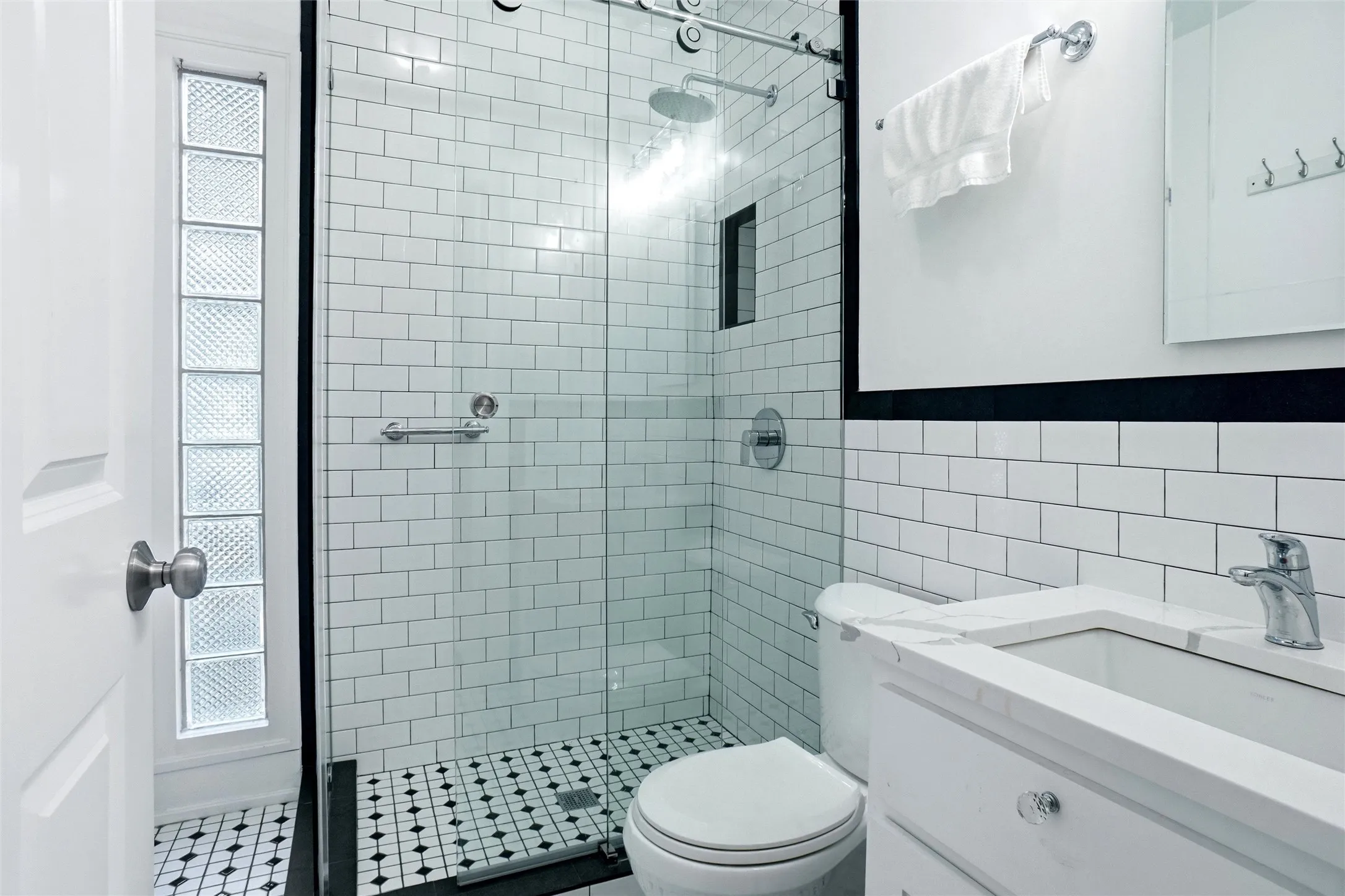 Bathroom featuring white subway tile, a glass-enclosed shower, black and white mosaic floor tile, a white vanity with a rectangular sink, and a glass block window