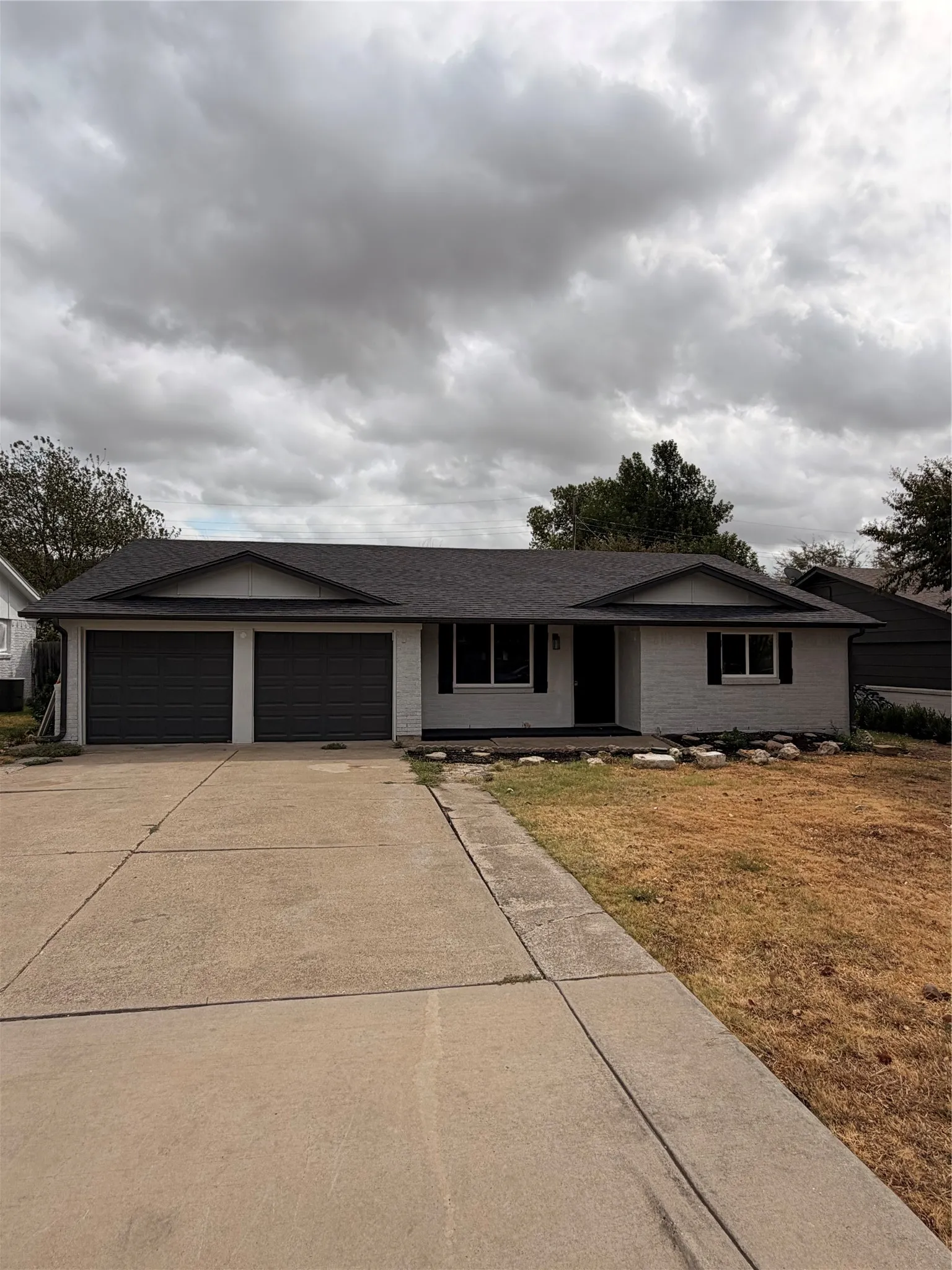 White brick exterior featuring a dark asphalt shingle roof, two-car garage with dark doors, and a concrete driveway