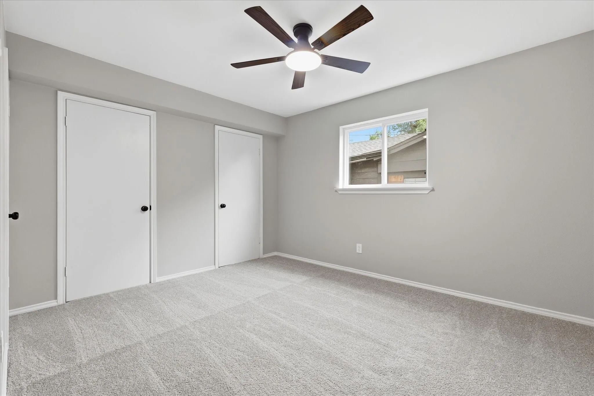 Spacious room featuring light gray wall paint, light-toned carpet flooring, a window with exterior views, and a ceiling fan with integrated lighting