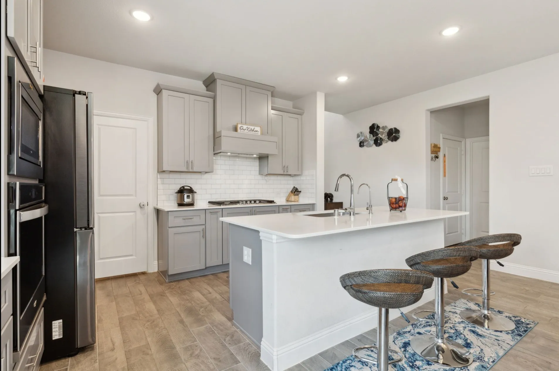Modern kitchen featuring light gray cabinetry, built-in stainless steel appliances, wood-finish flooring, white subway tile backsplash, and a central island with a sink and white countertop