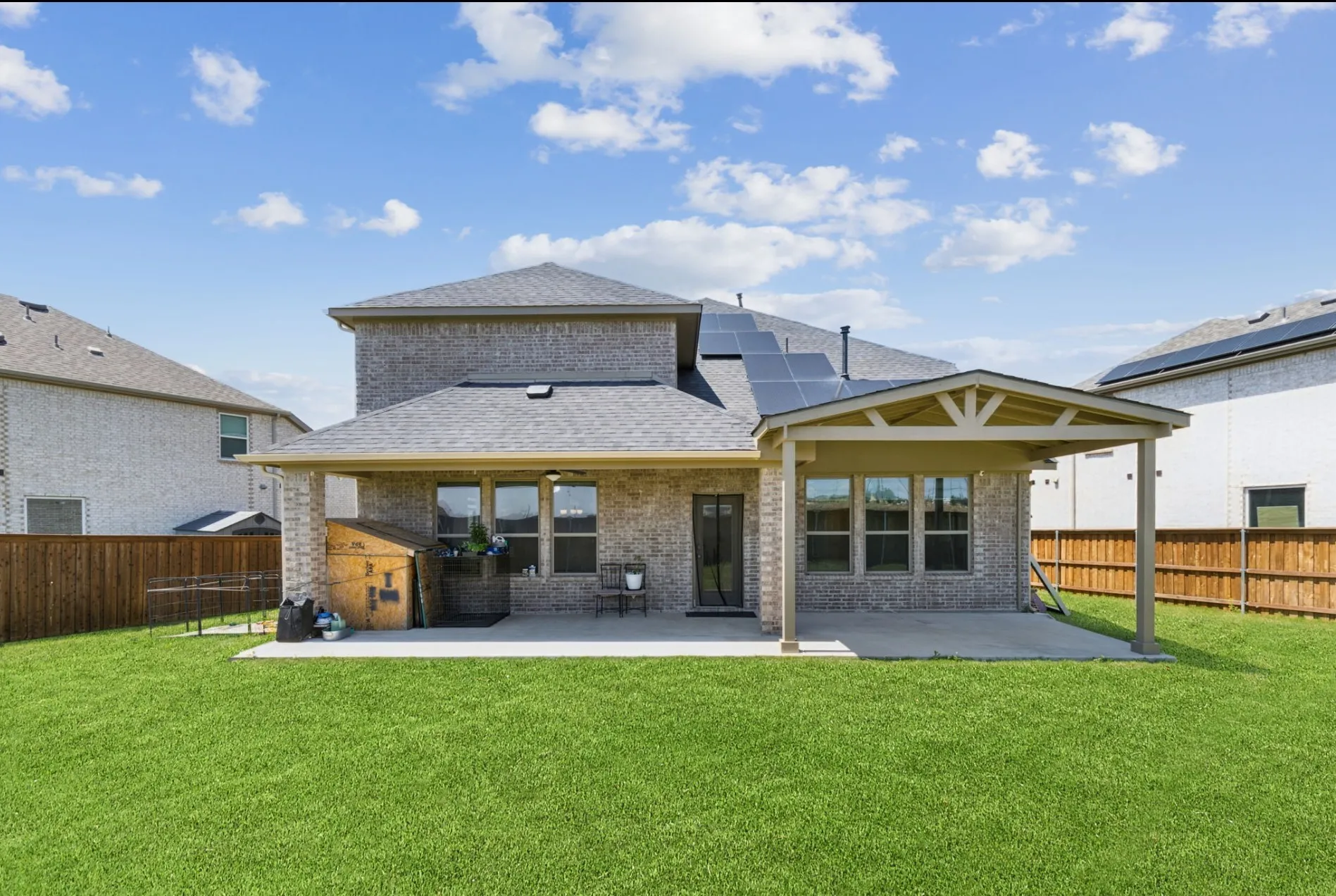 Expansive green lawn with a covered patio, brick exterior, dark grey shingle roof, and solar panels