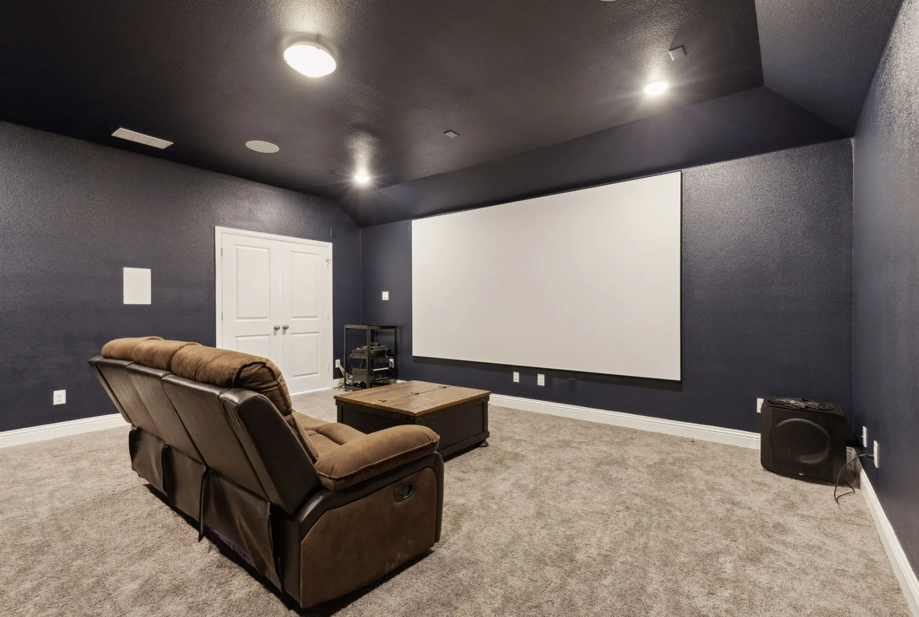 Dedicated media room featuring a large projection screen, dark painted walls, recessed lighting, and a textured carpeted floor