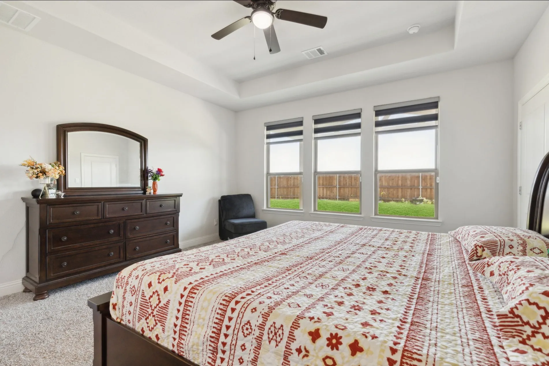 Bedroom featuring a tray ceiling with a ceiling fan, three horizontal windows with roller shades, and light gray carpet flooring