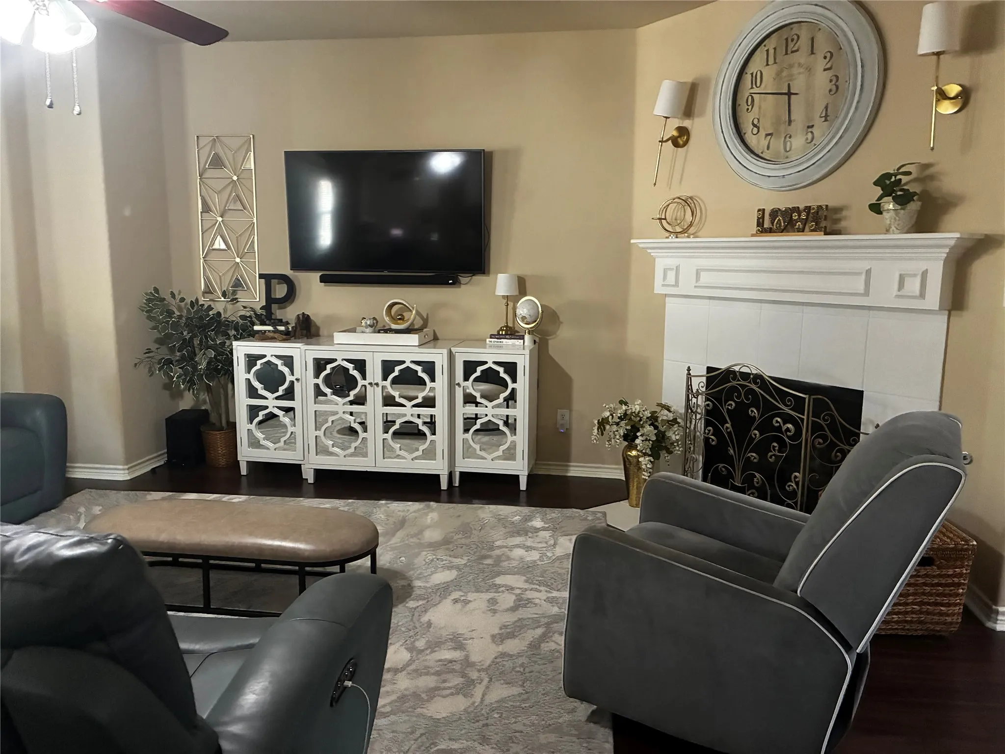 Living area featuring rich wood-finish flooring, a white mantelpiece above a fireplace with an ornate screen, and a ceiling fan