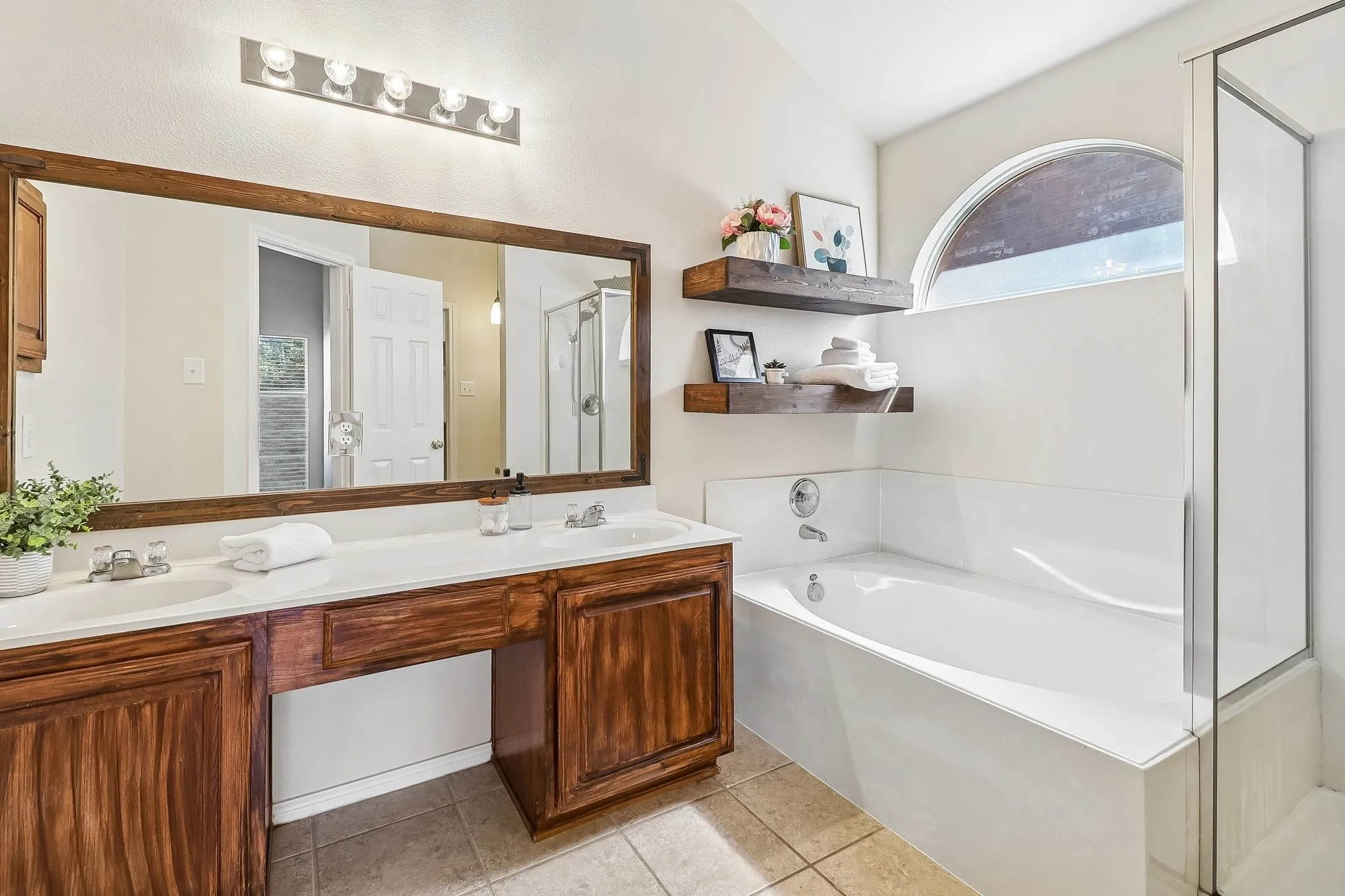 Bathroom featuring a spacious double vanity with wood cabinetry, a large framed mirror, and a built-in bathtub with an arched window