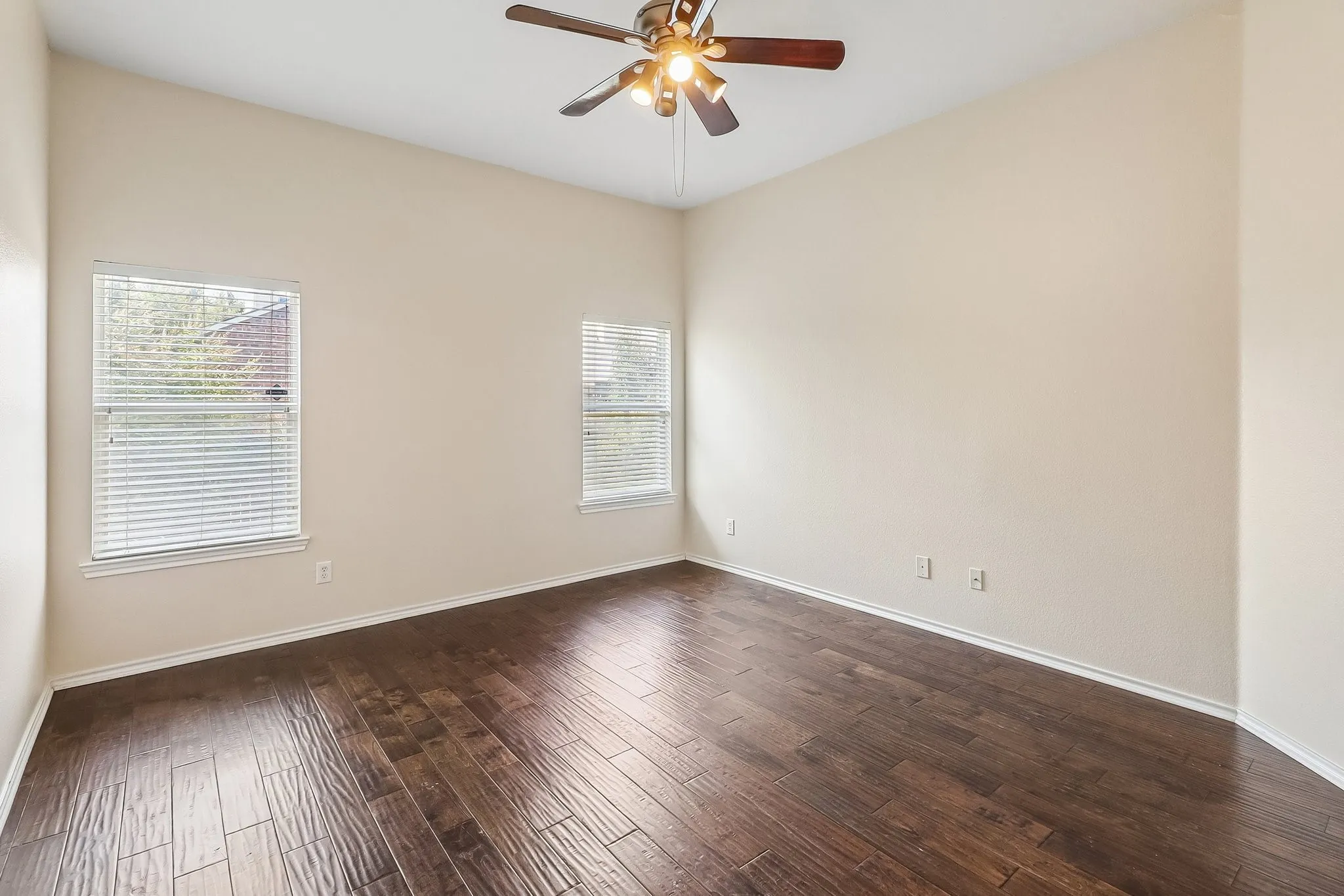 Spacious room featuring rich wood-finish flooring, two windows with blinds, a ceiling fan with integrated lighting, and neutral wall tones