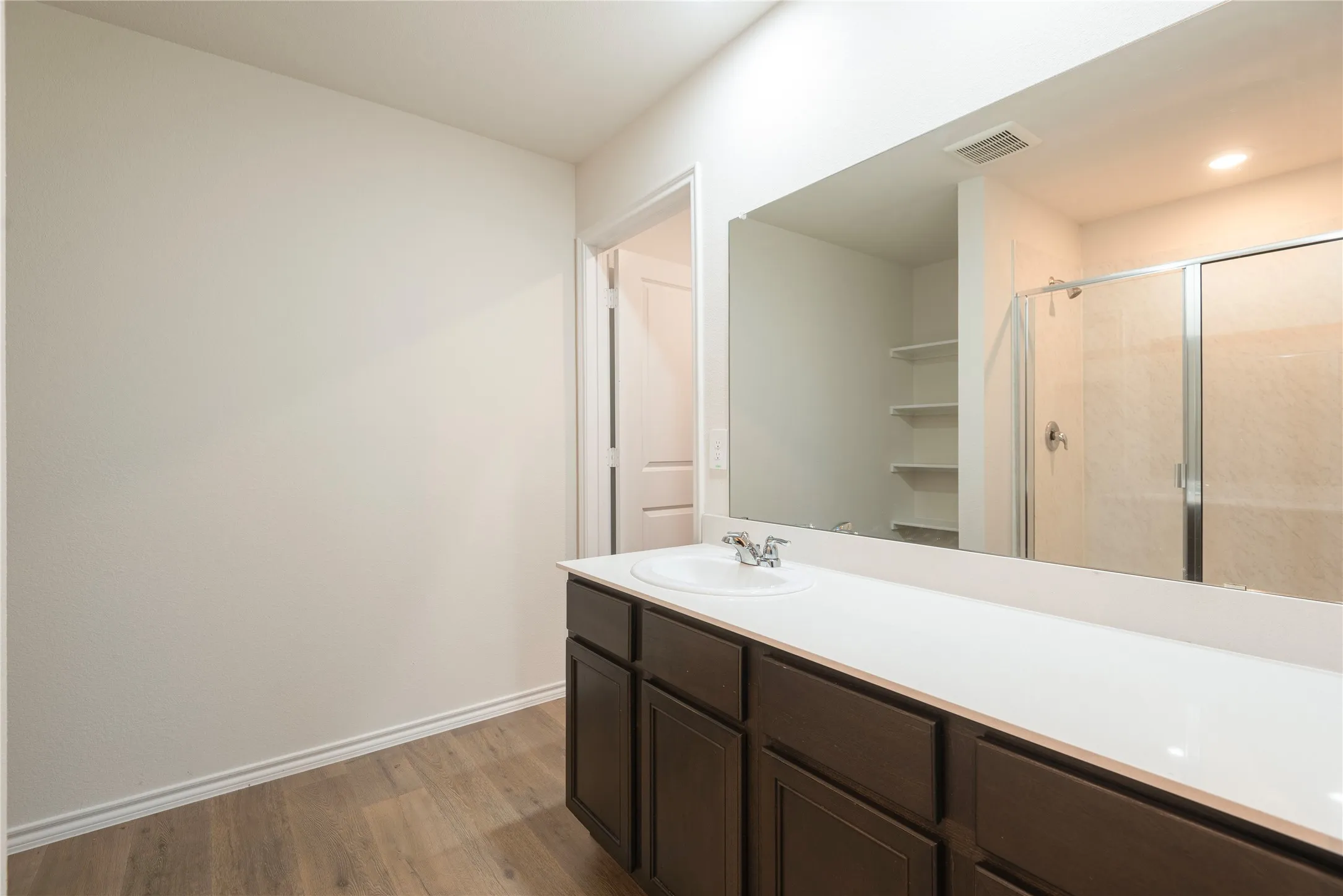 Bathroom featuring wood-finish flooring, a white countertop vanity with dark cabinetry, a large wall-mounted mirror, and a glass-enclosed shower