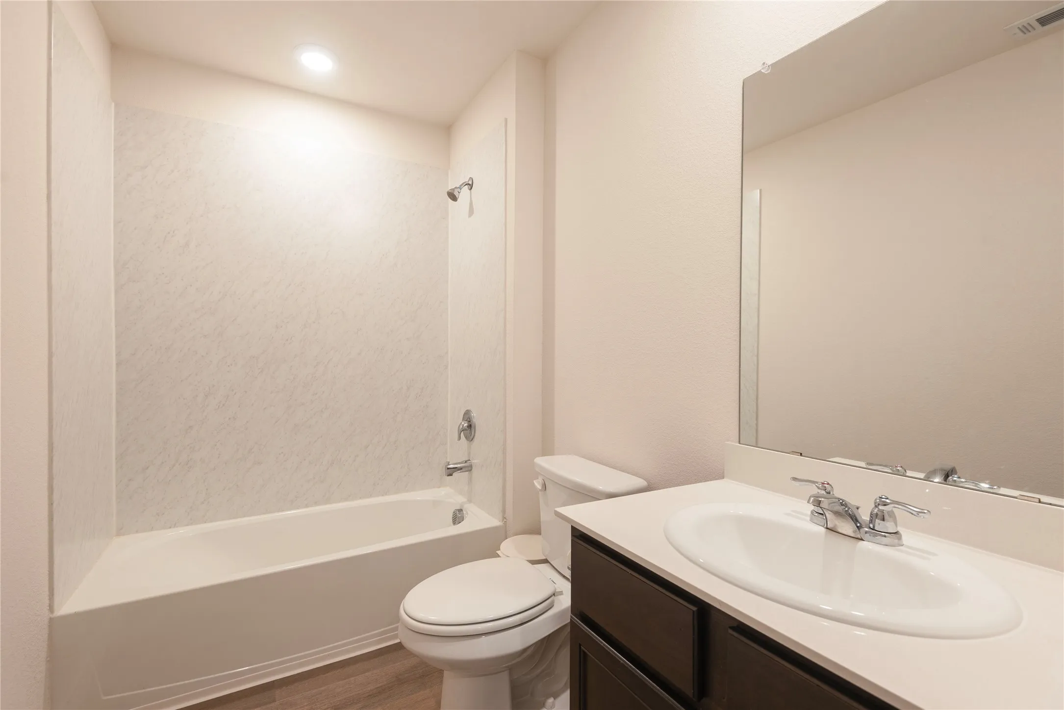 Full bathroom featuring a white bathtub/shower combination with a textured surround, a dark wood-finish vanity with a white countertop and integrated sink, a large wall-mounted mirror, and wood-finish flooring