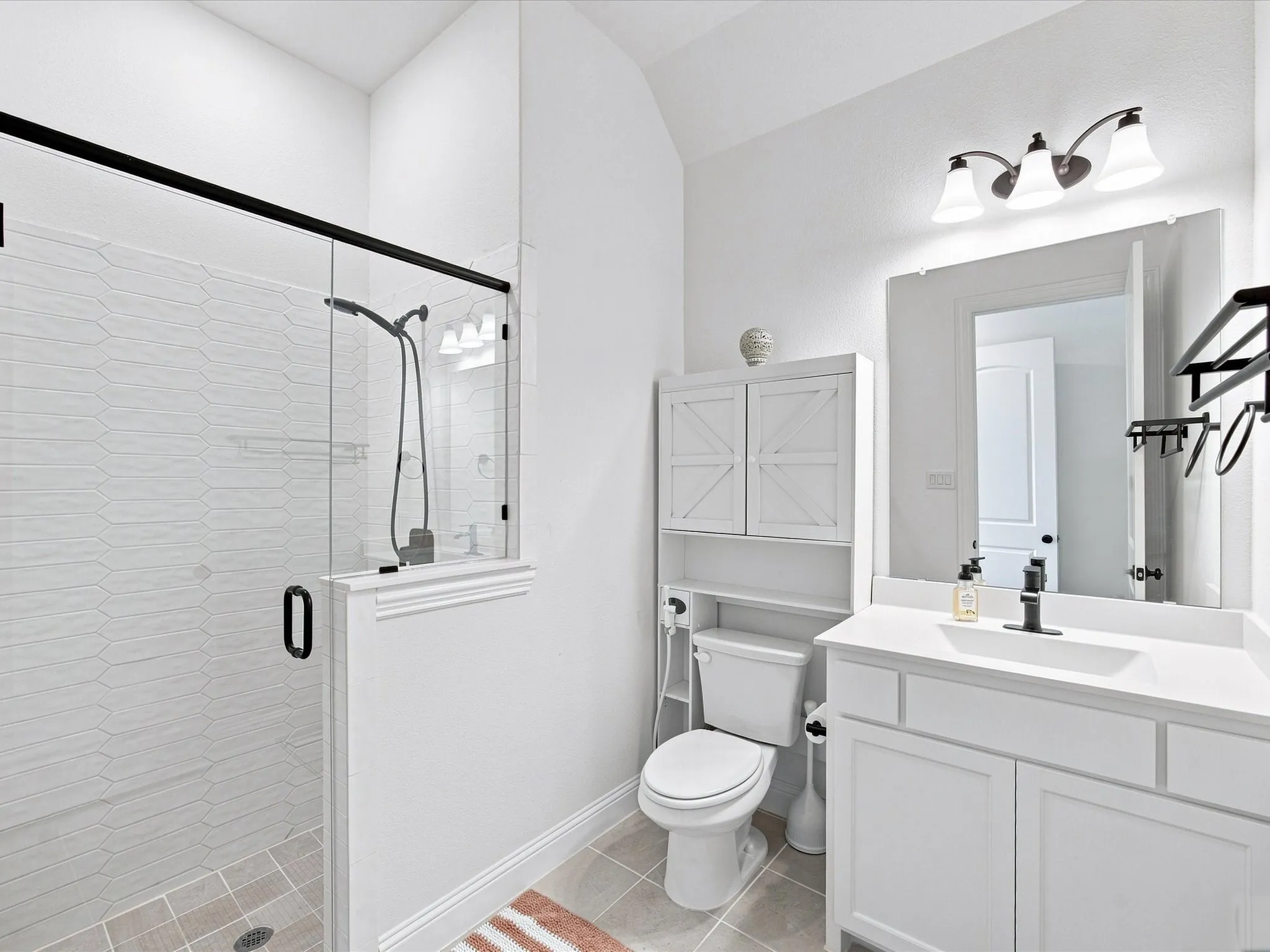 Glass-enclosed shower with elongated hexagon tile, white vanity with integrated sink, sleek matte black fixtures, and white over-the-toilet storage cabinetry