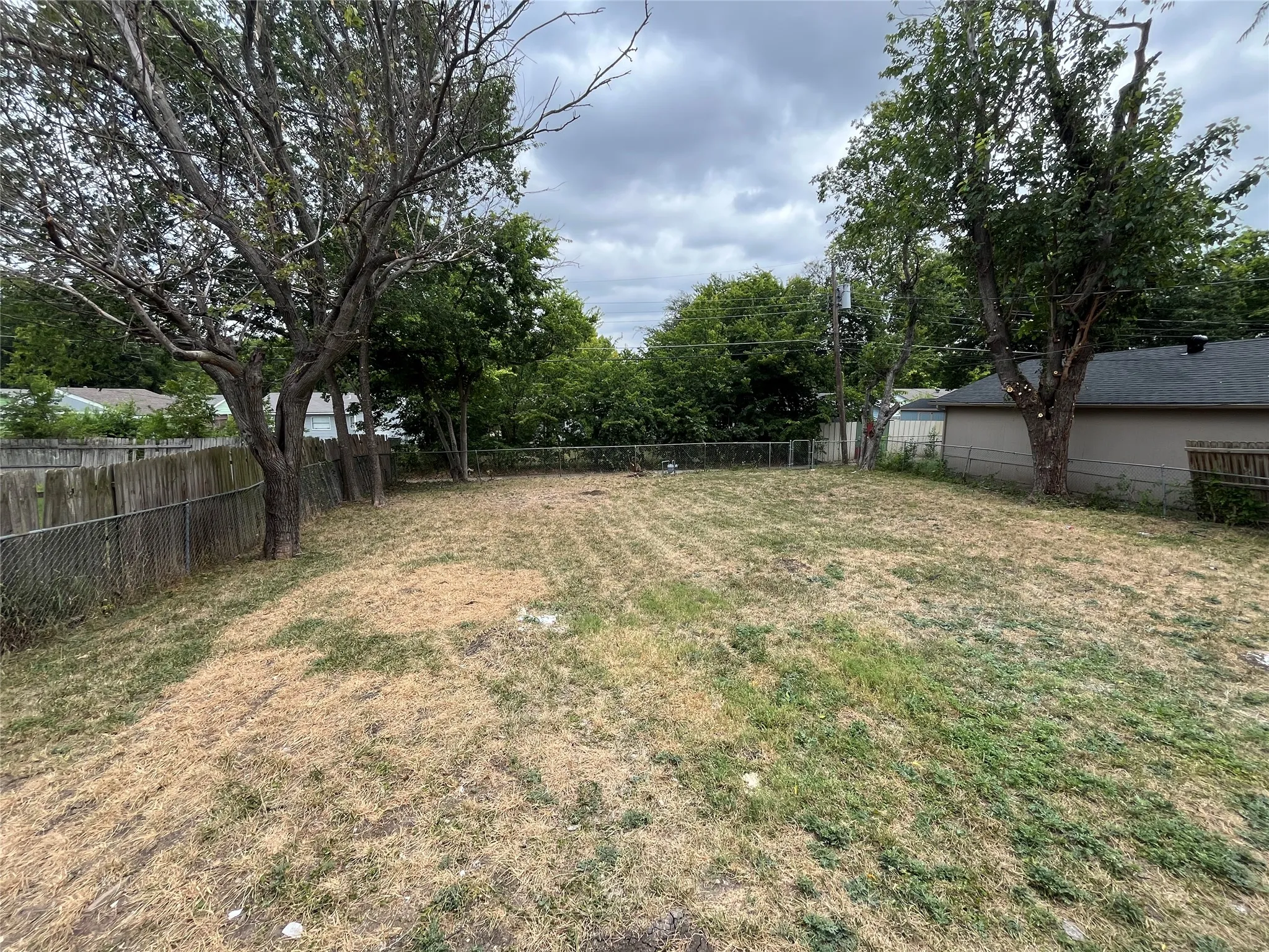 Expansive rear yard featuring a chain-link fence, mature trees, and a combination of natural ground cover and grass