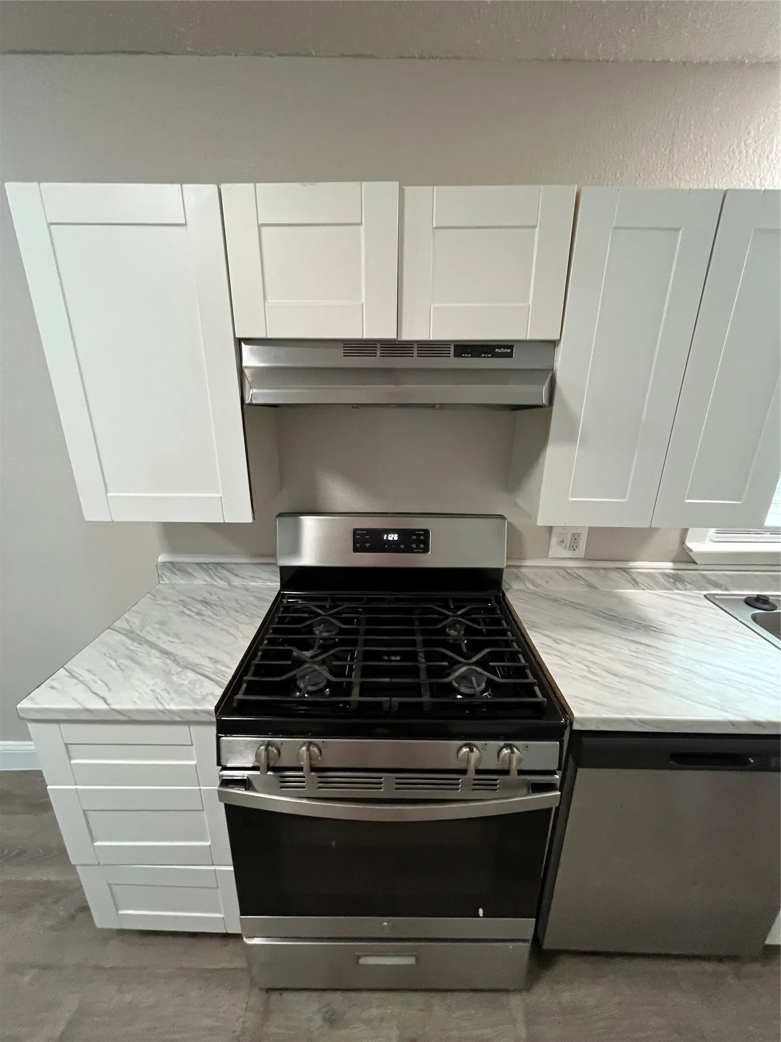 Stainless steel range with a gas cooktop, under-cabinet range hood, white shaker-style cabinetry, white countertops with gray veining, and wood-finish flooring