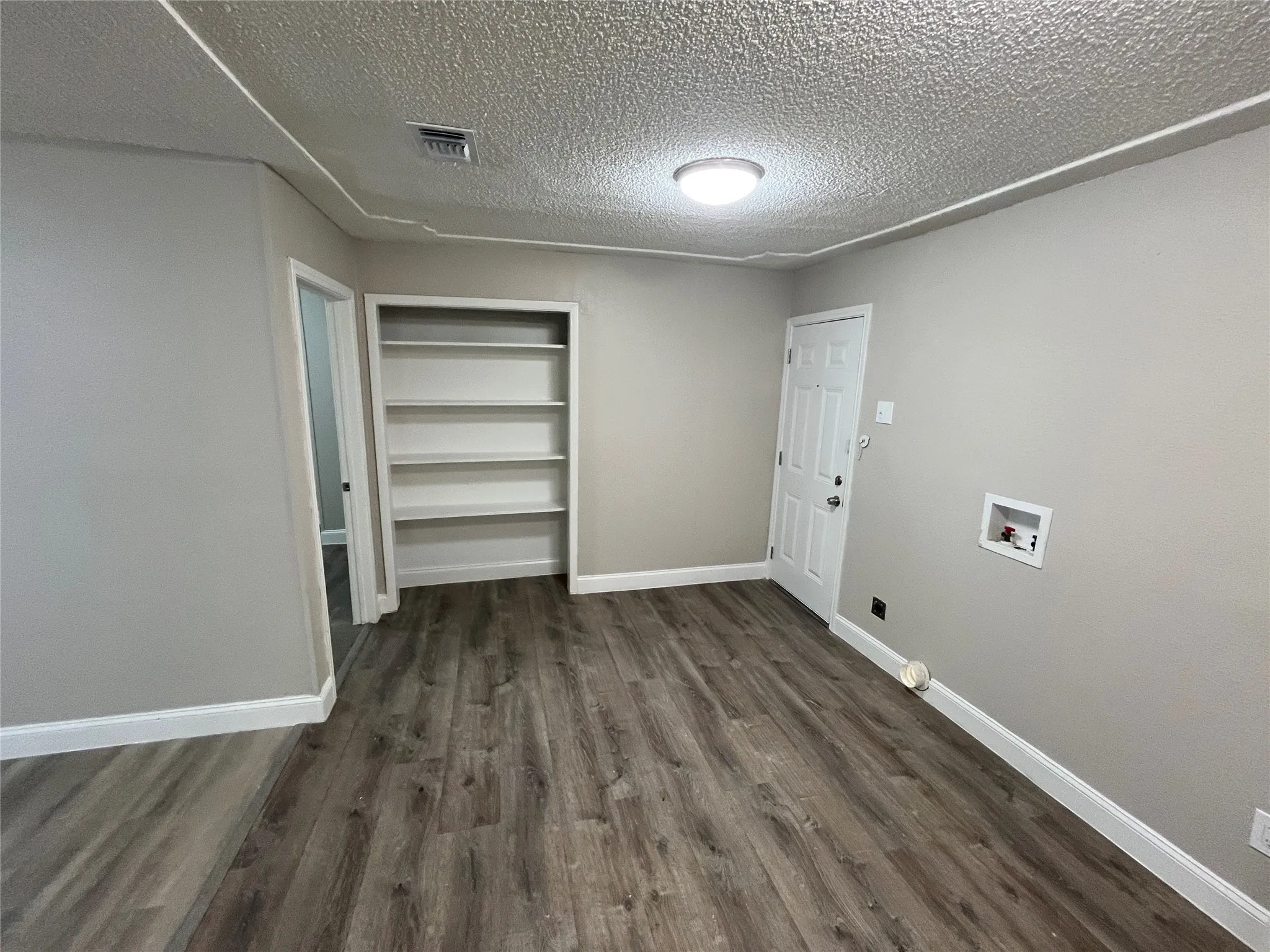 Room featuring neutral wall paint, wood-finish flooring, built-in shelving, a paneled door, and a utility hookup