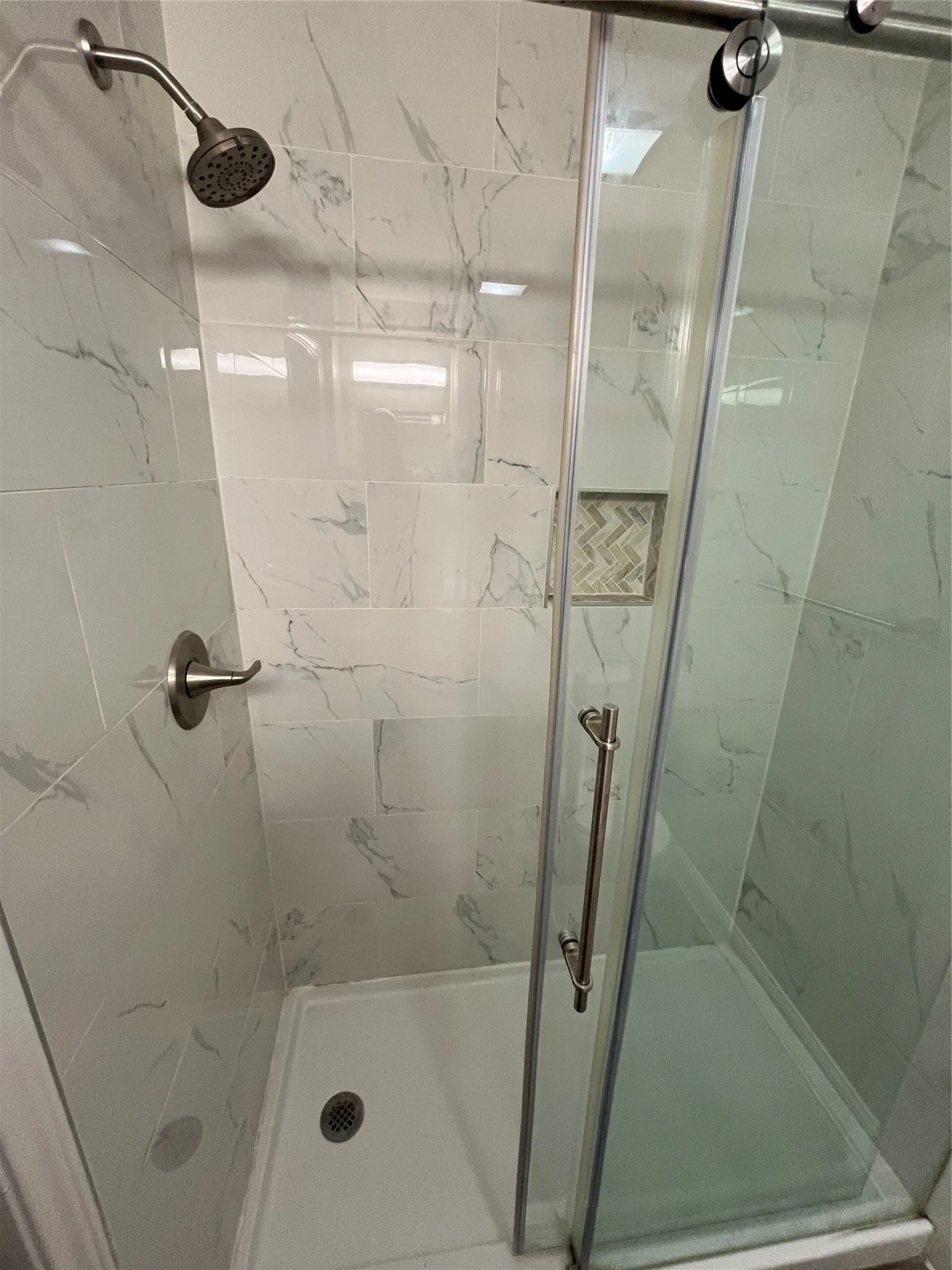 Shower enclosure featuring white marble-look subway tile, a recessed niche with herringbone pattern accent tile, a sliding glass door, a white shower pan, and brushed nickel fixtures