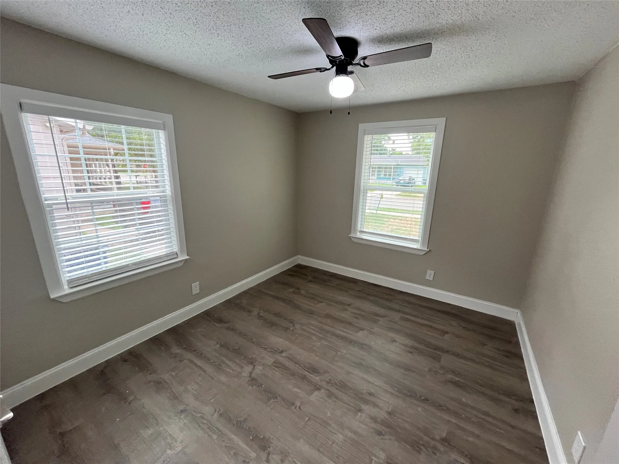 Room featuring wood-finish flooring, two windows with white blinds, and a dark-toned ceiling fan with an integrated light fixture