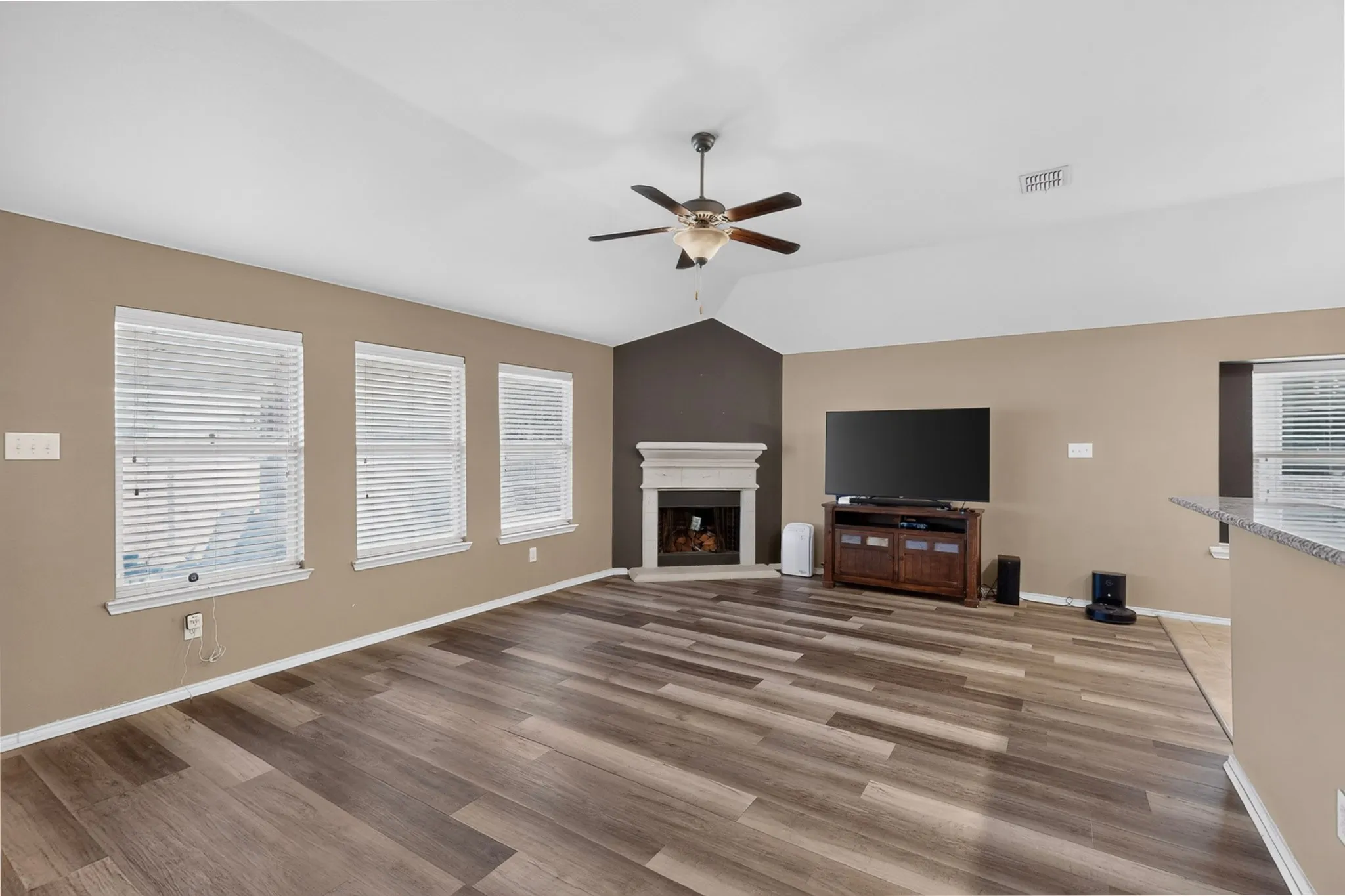 Living area featuring wood-finish flooring, a vaulted ceiling, a ceiling fan, a fireplace with a mantel, and multiple windows with blinds