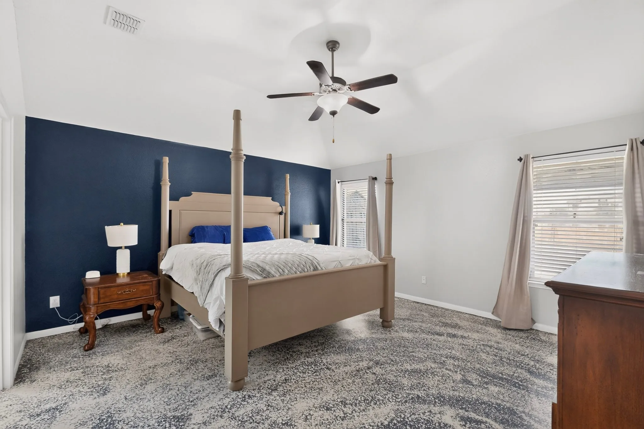 Vaulted ceiling, ceiling fan with light fixture, large window with blinds, accent wall, and patterned carpeting