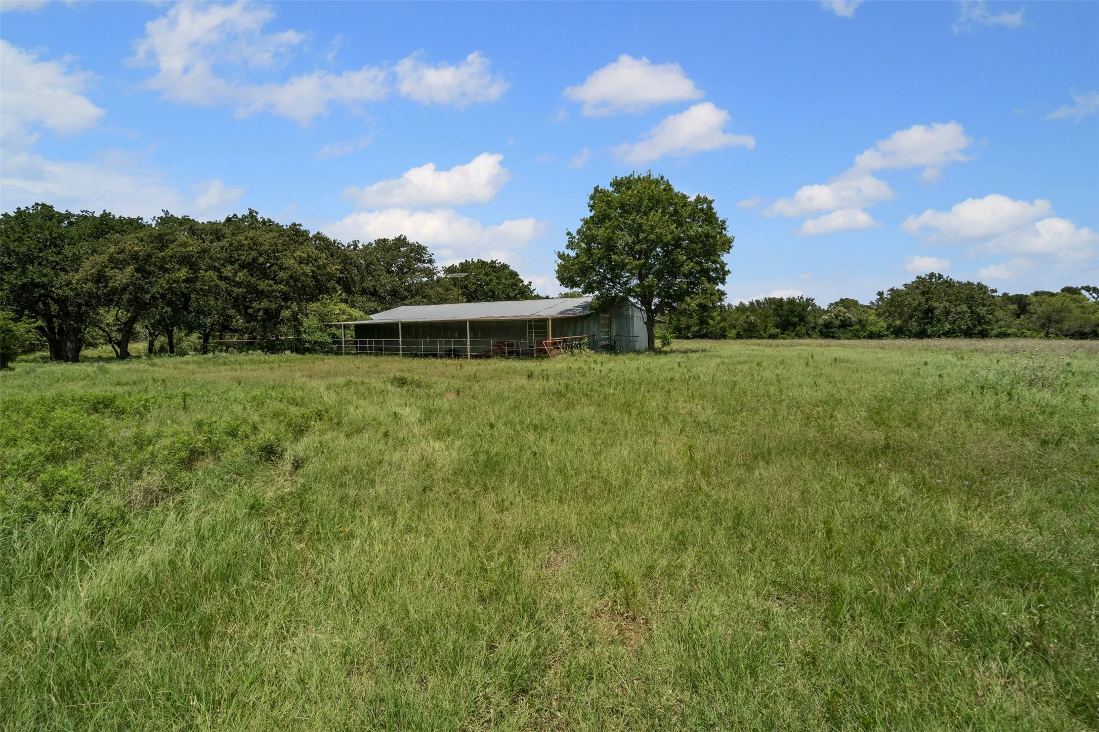 Expansive green field featuring a large barn with a metal roof and an open-air side, bordered by mature trees