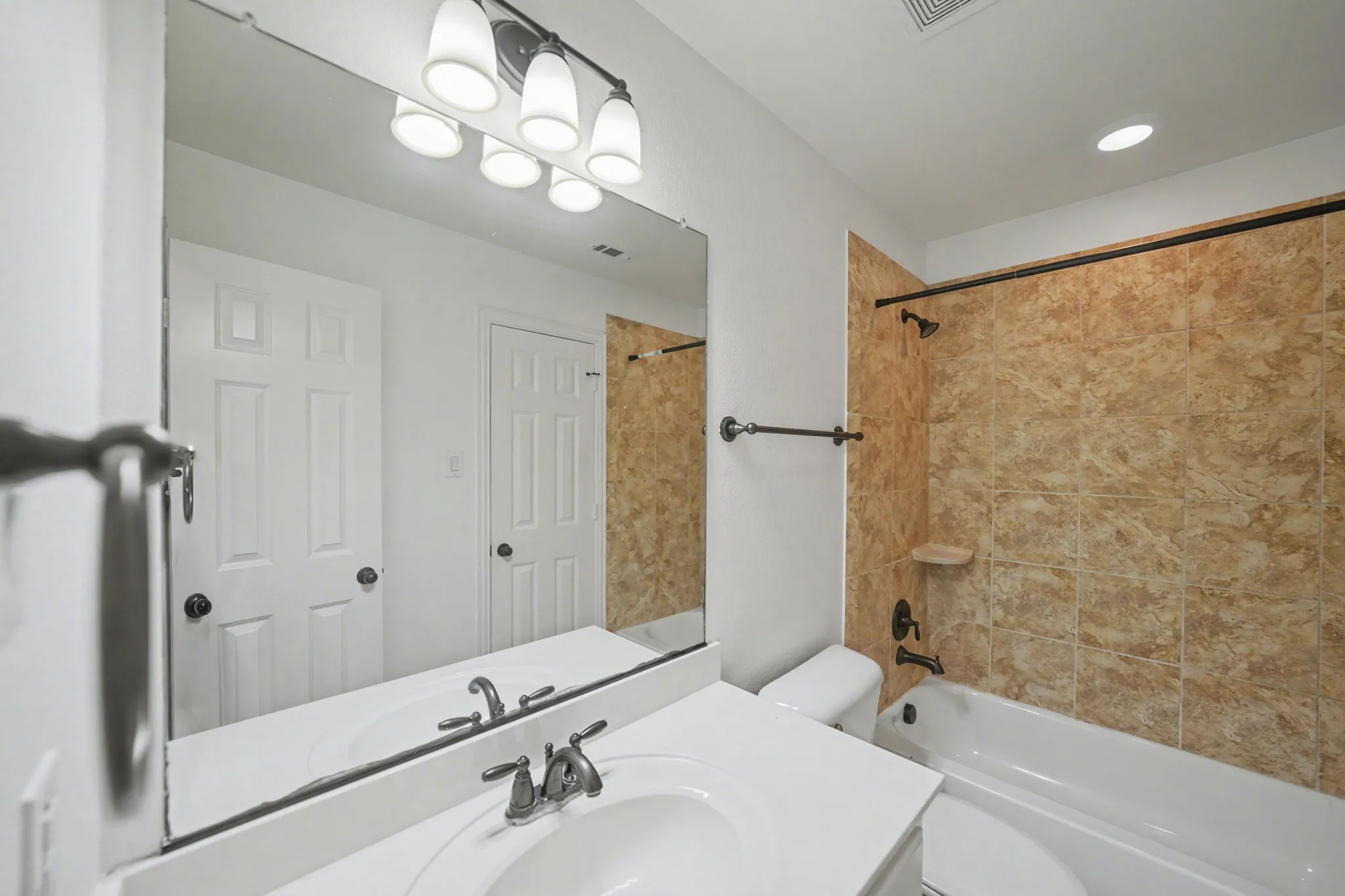 Bathroom featuring a vanity with a white countertop and integrated sink, an overhead light fixture with four white glass shades, a full-width mirror, a bathtub with a tiled surround, and dark-finish hardware