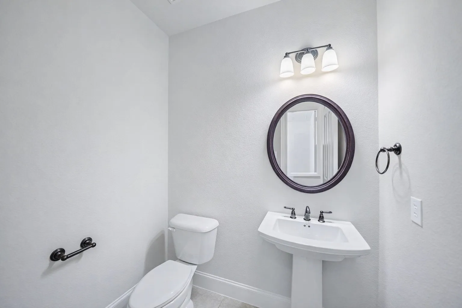 Pedestal sink with a two-handle faucet, circular framed mirror, and a three-light vanity fixture