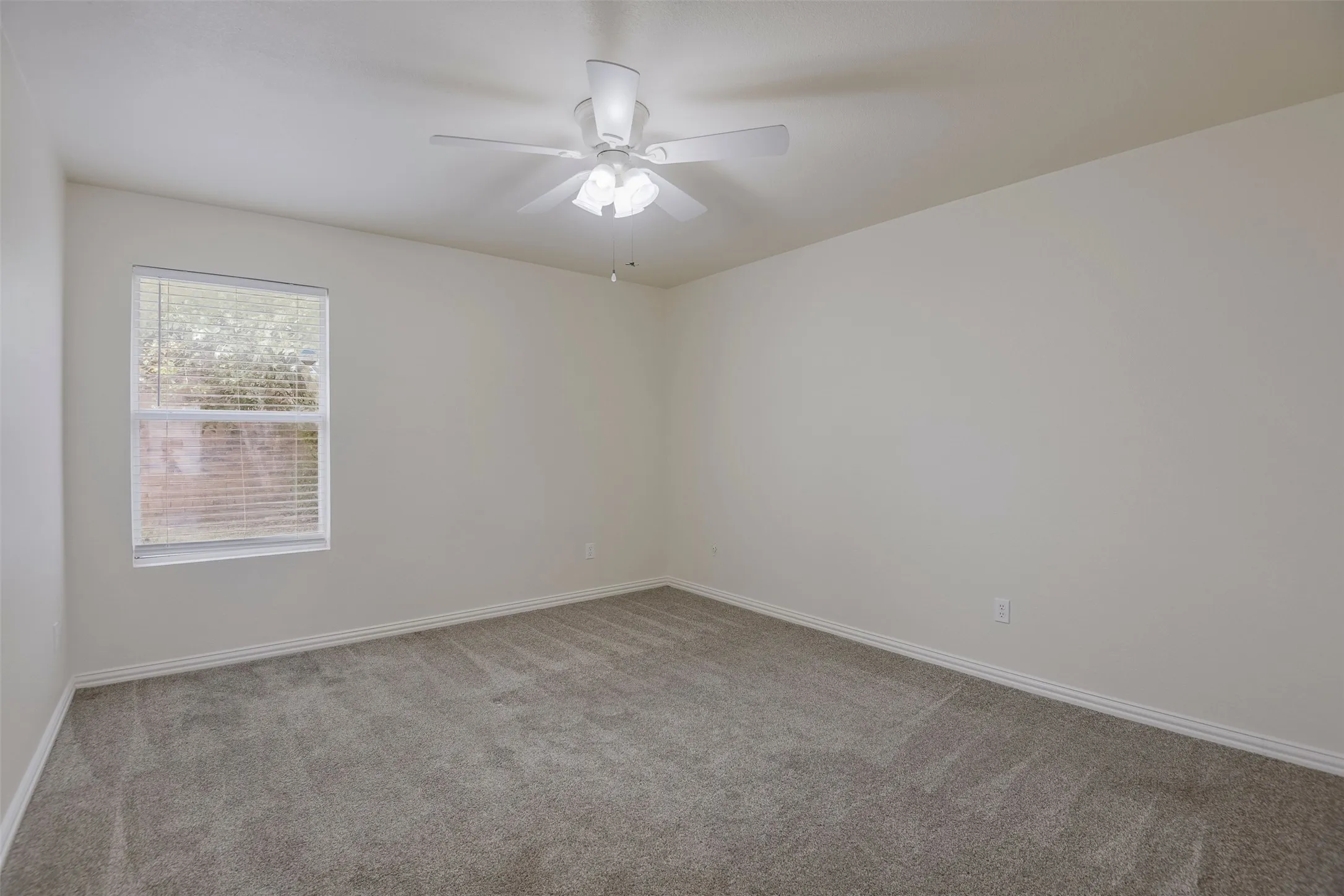 Spacious room featuring neutral carpeting, a ceiling fan with light fixture, and a window with blinds
