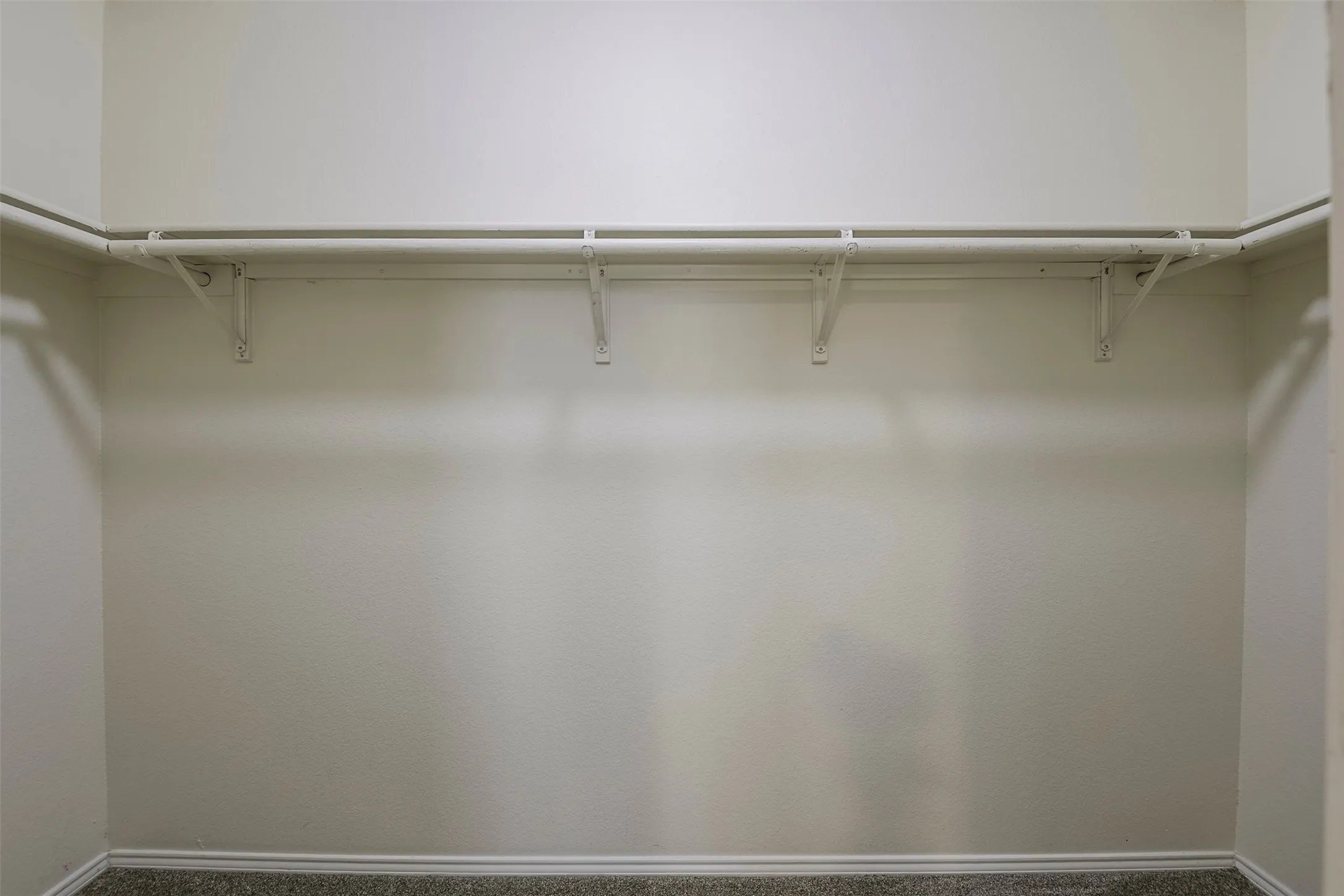 Closet featuring a single clothes rod, wall-mounted shelving brackets, light neutral wall paint, and gray carpet flooring with baseboards
