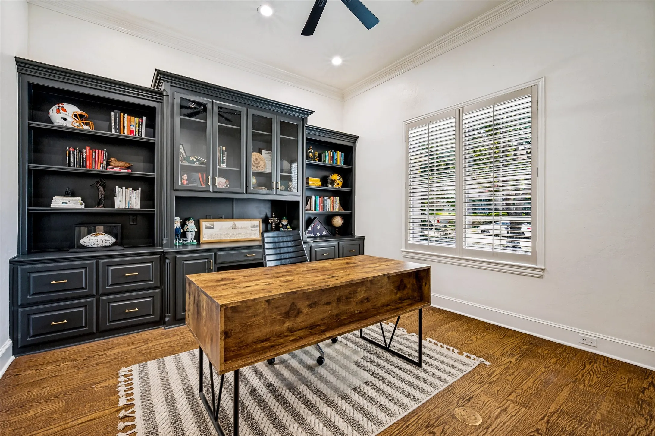 Built-in cabinetry with glass-front uppers and lower drawers, wood-finish flooring, crown molding, and a window with plantation shutters