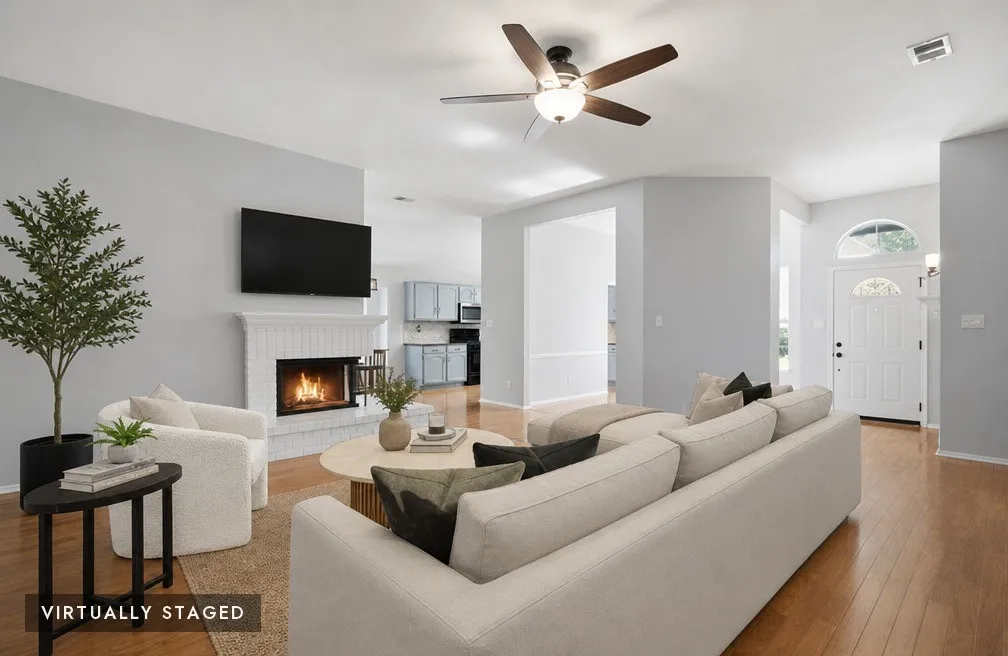 Spacious living area featuring light wood-finish flooring, a white brick fireplace with a mantel, and a ceiling fan with integrated lighting, virtually staged