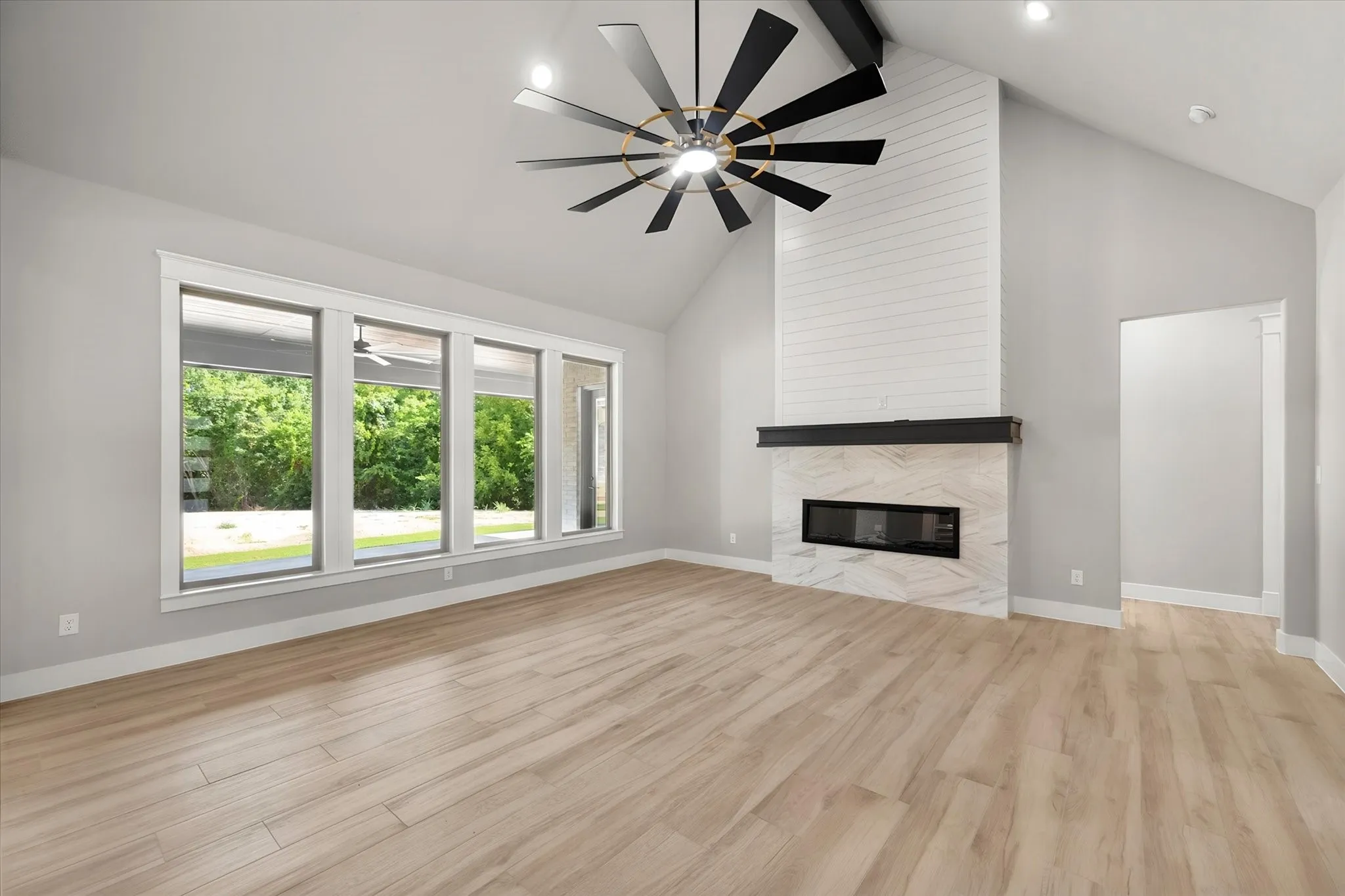 Vaulted ceiling with exposed beam, large picture windows, wood-finish flooring, fireplace with stone surround and shiplap accent, and a contemporary ceiling fan