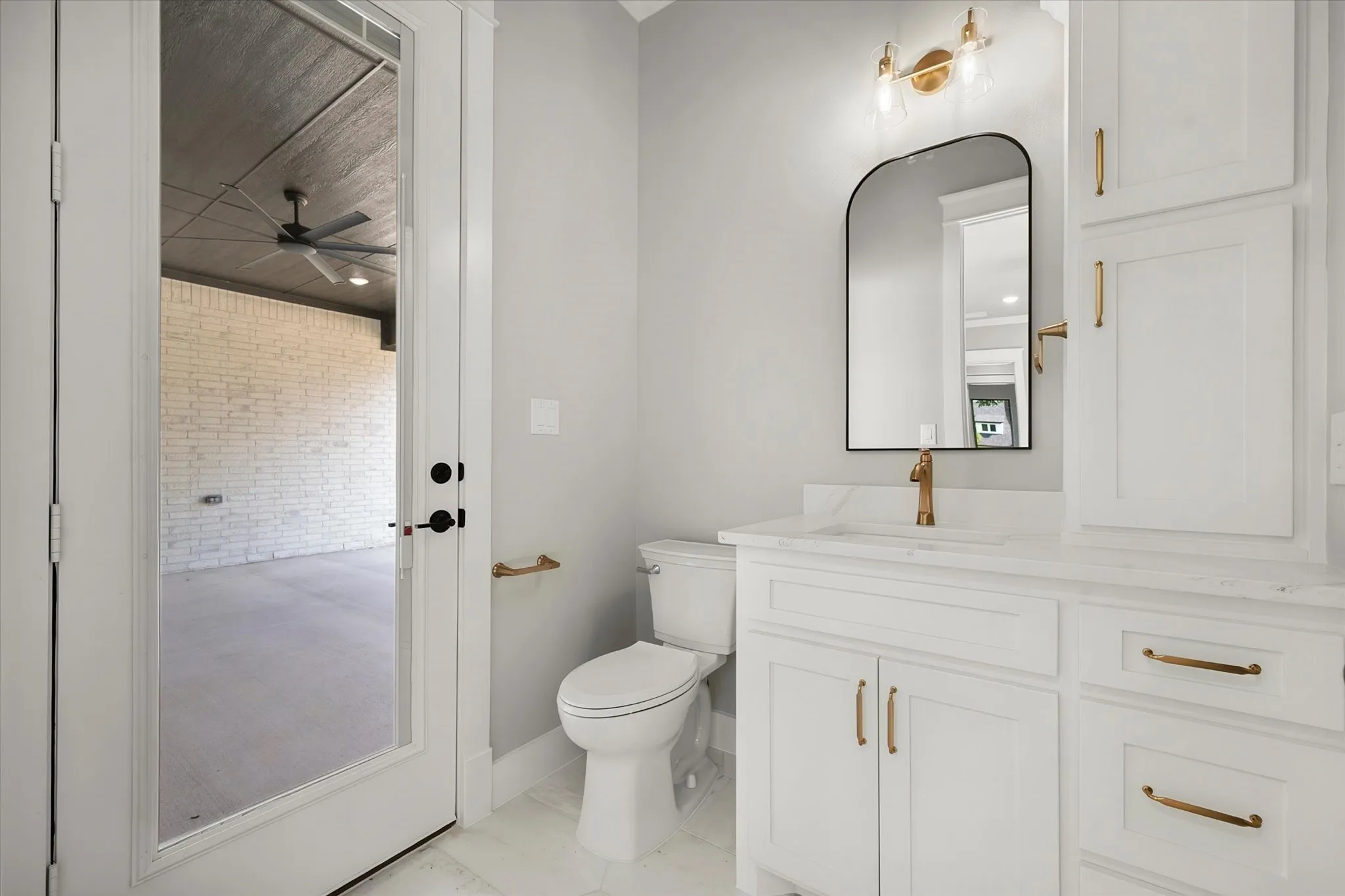 White shaker cabinetry with gold-tone hardware, a white countertop with an integrated sink, a contemporary arched mirror, and a gold-tone wall sconce