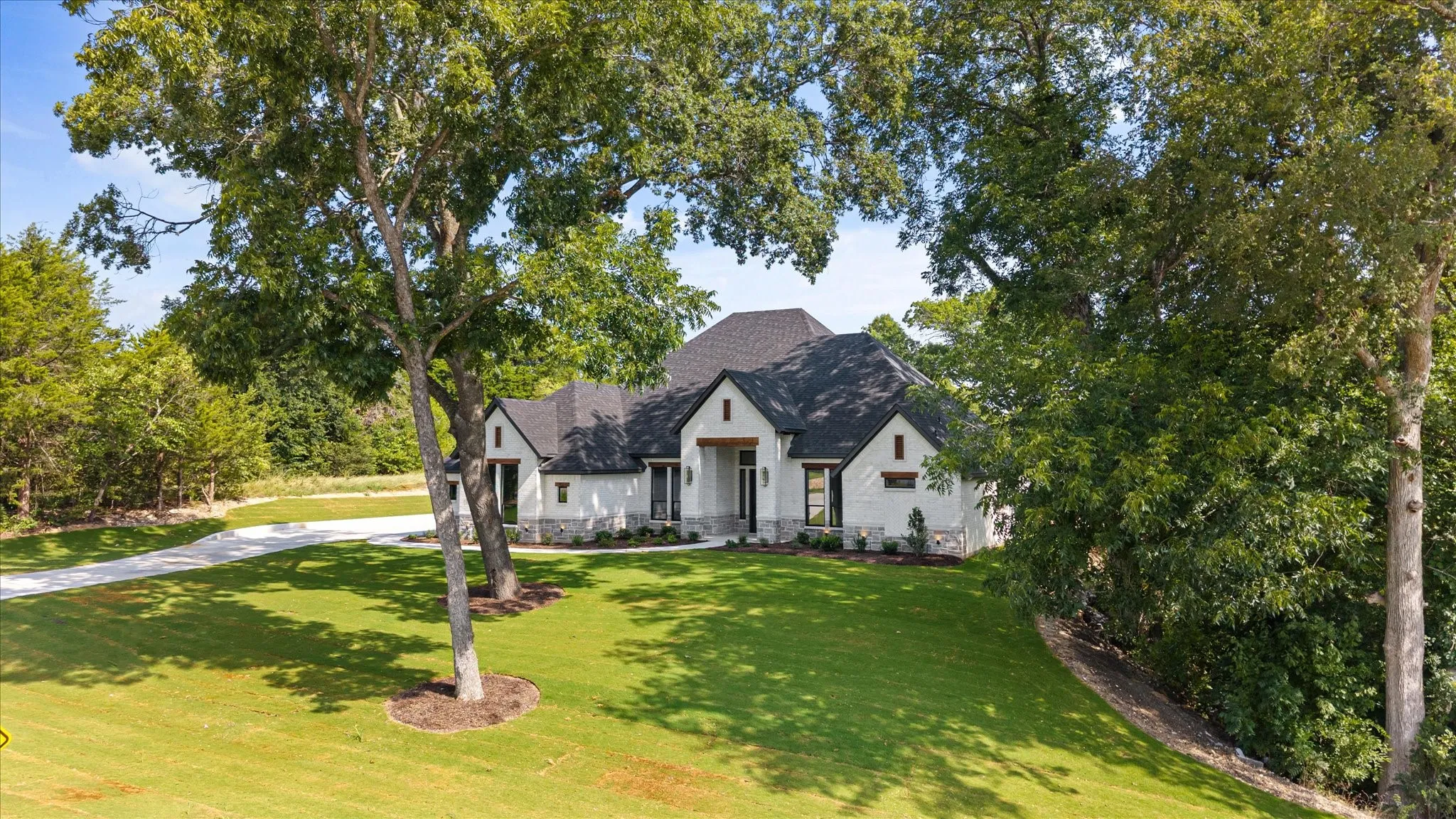 Brick and stone exterior with a dark shingle roof, a paved driveway, and mature landscaping