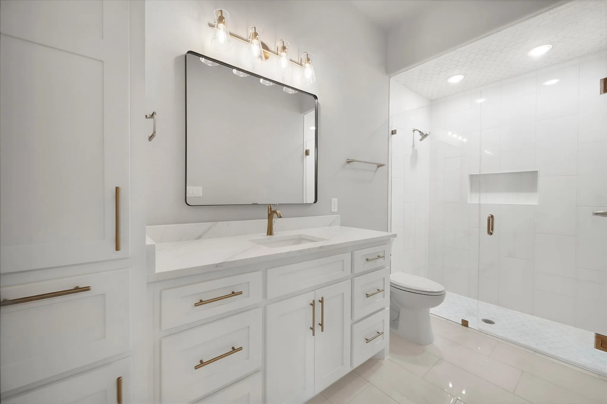 White shaker-style cabinetry with gold-tone hardware, white countertop with integrated sink, gold-tone single-handle faucet, frameless shower enclosure, and white tile flooring