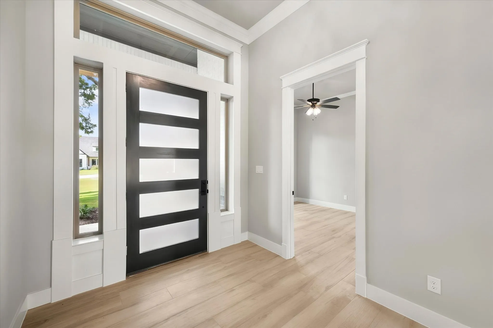 Modern foyer showcasing a contemporary black horizontal-slat entry door with sidelite window, light wood-finish flooring throughout, white painted walls and trim, recessed ceiling detail, and ceiling fan fixture